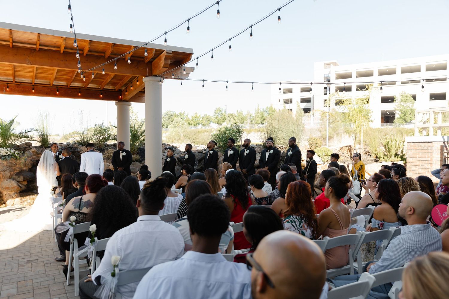 Outdoor event space filled with guests seated at tables under string lights and wooden pergola during daytime gathering.