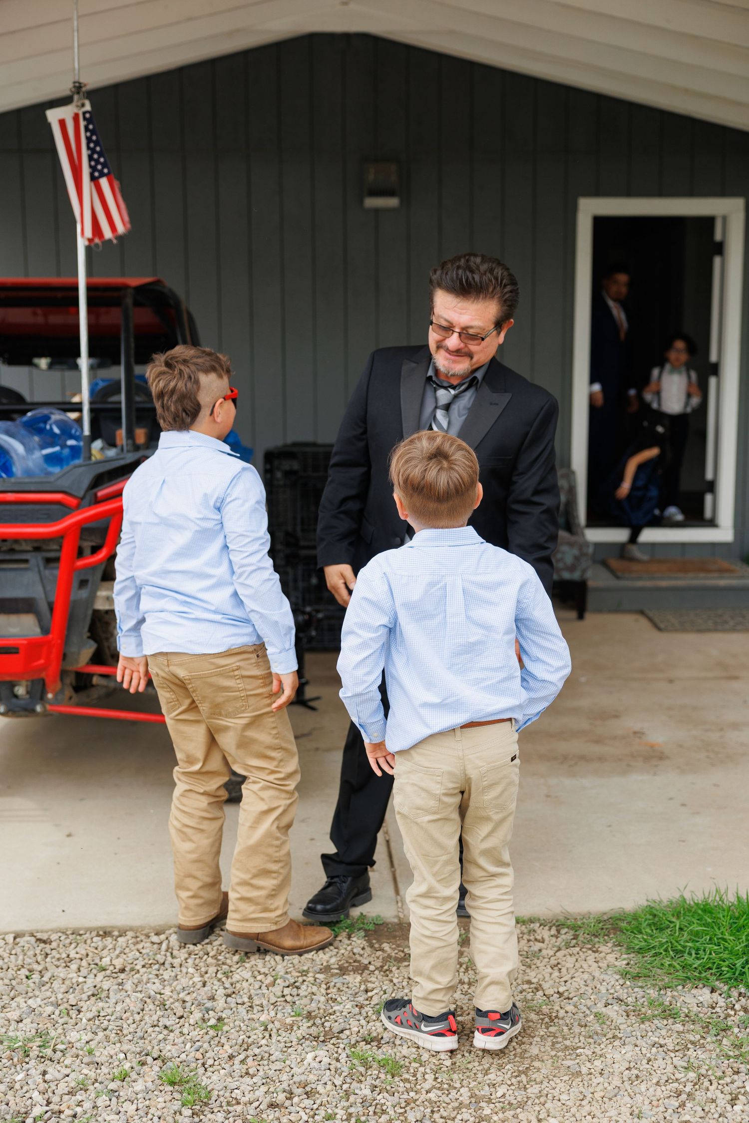 Two young boys in matching light blue shirts and khakis stand with an adult in a suit outside a garage with an American flag.