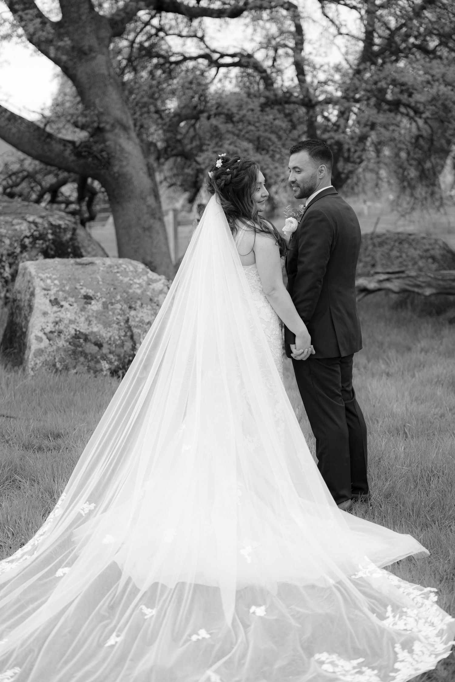 A black and white wedding photo showing a couple in formal attire with a flowing cathedral veil in a rustic outdoor setting.