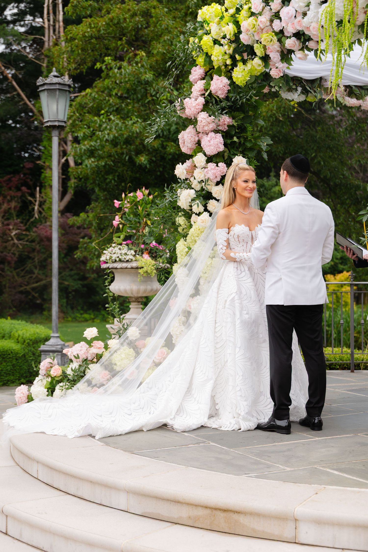 A couple in wedding attire poses for photographs on elegant stone steps surrounded by pink and yellow floral arrangements.