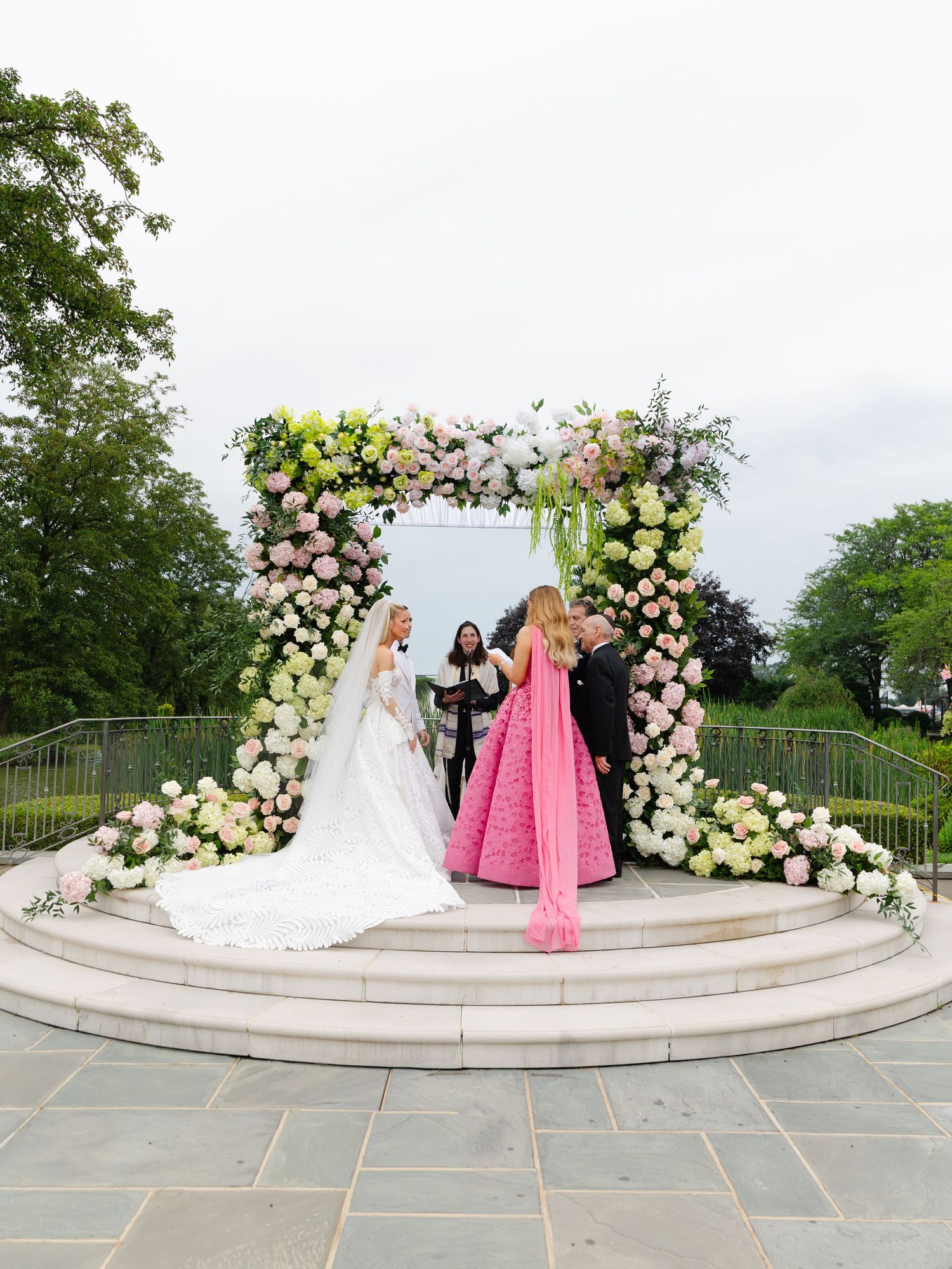 Wedding ceremony under an elegant floral arch with white and pink roses on a curved stone terrace overlooking a garden.