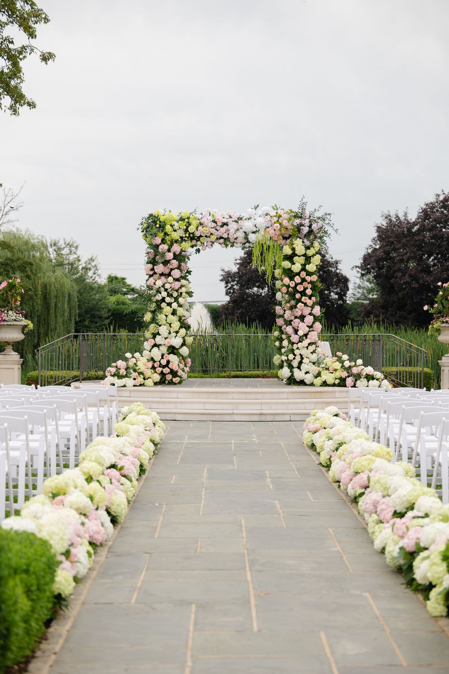 Elegant outdoor wedding ceremony setup with white chairs and lush floral arrangements lining the aisle against a garden backdrop.