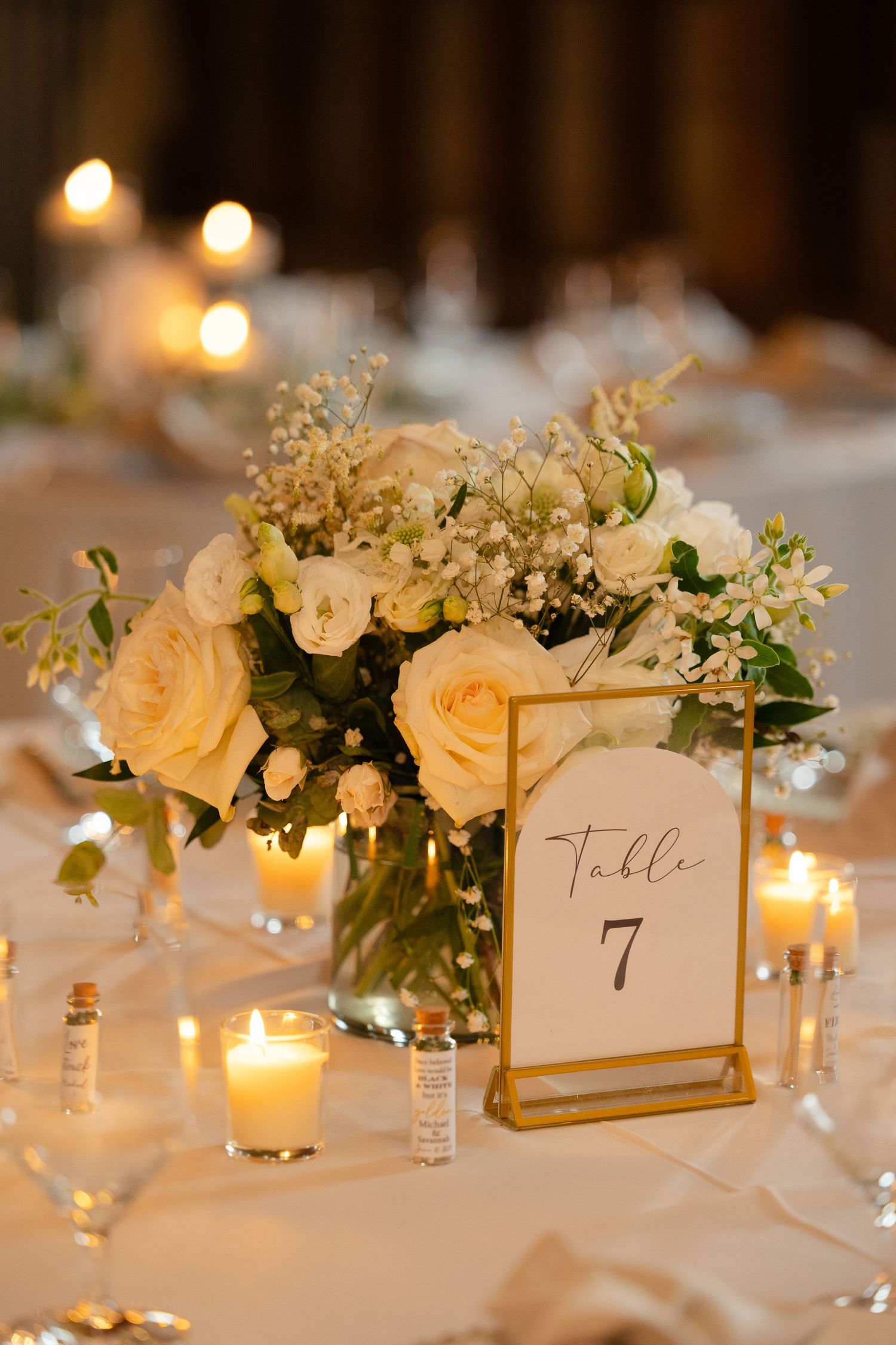Elegant wedding table centerpiece with peach roses and baby's breath next to a gold-framed table number and glowing candles.