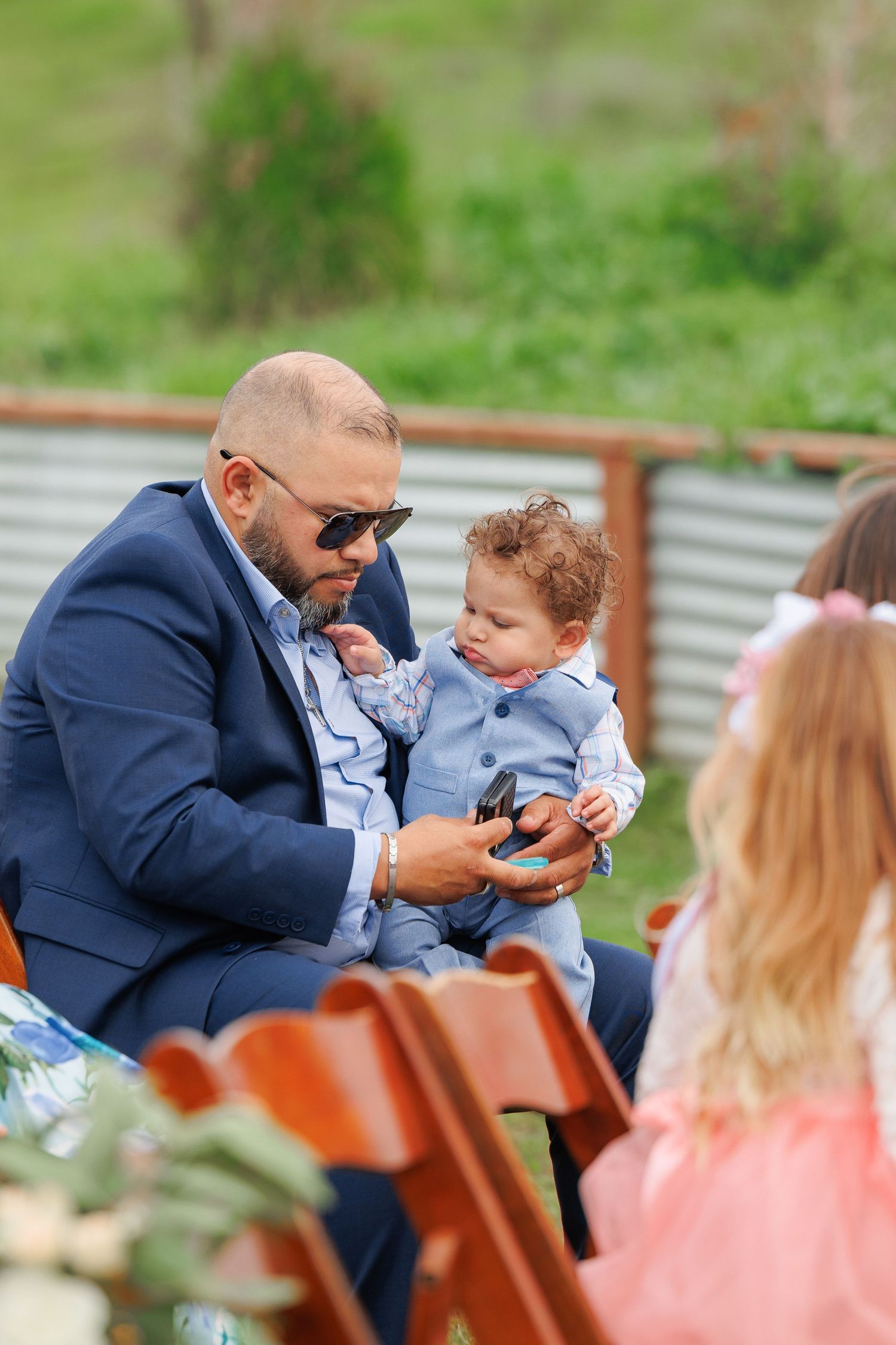 A person in a navy suit sits outdoors interacting with a small child during what appears to be a social gathering.