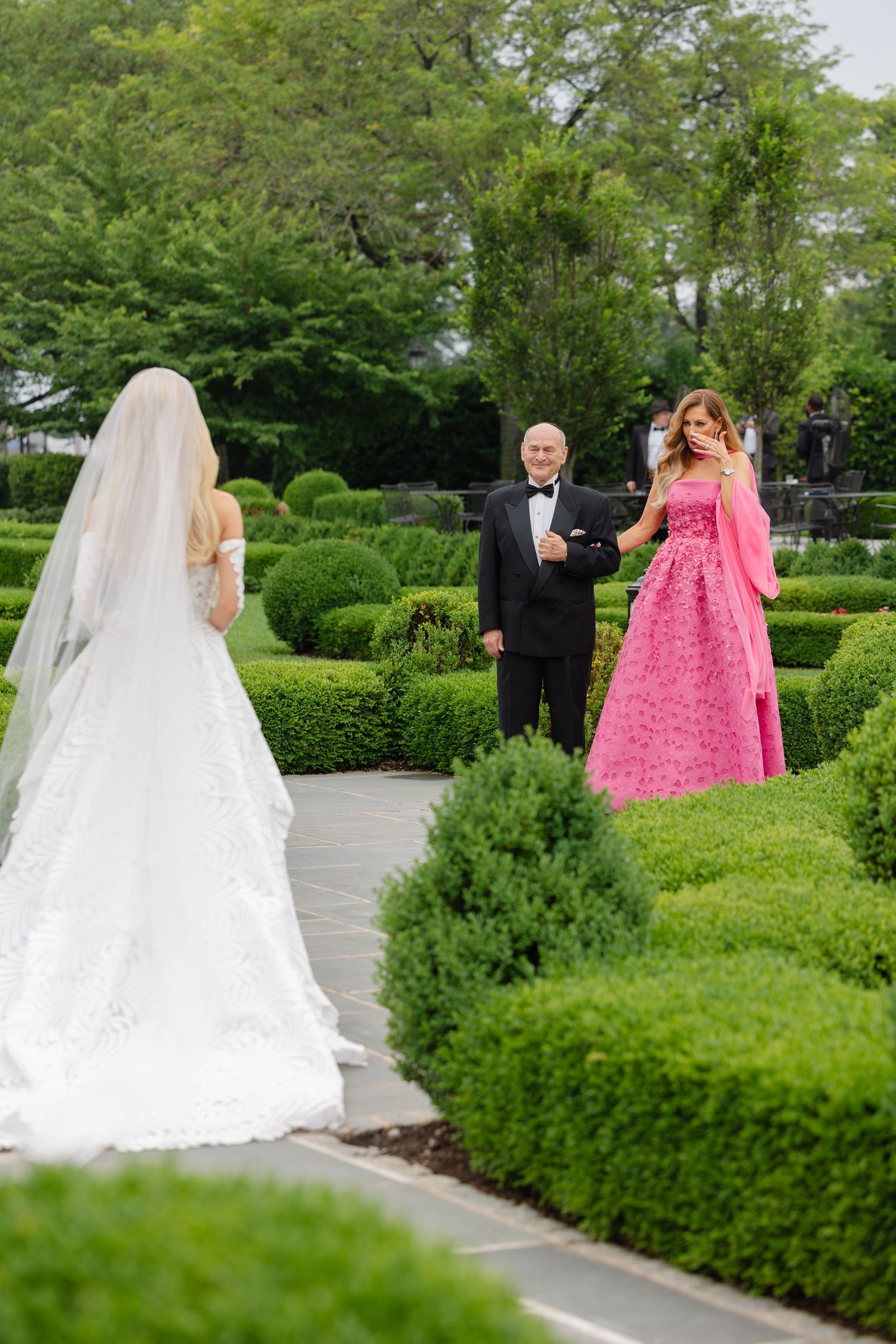 A wedding party walks through a lush garden path with manicured hedges and greenery on a sunny day.