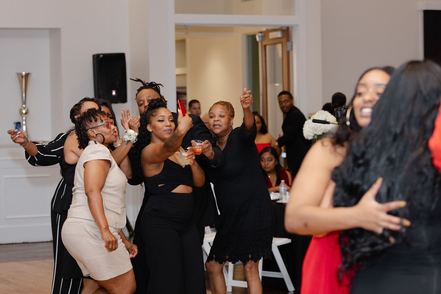 People dancing and socializing at an indoor party or celebration event with white walls and dark attire.