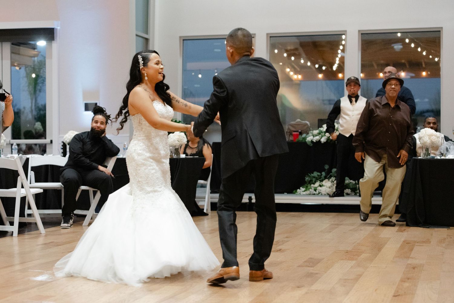 Wedding guests dance and celebrate on the dance floor during an indoor reception with string lights overhead.