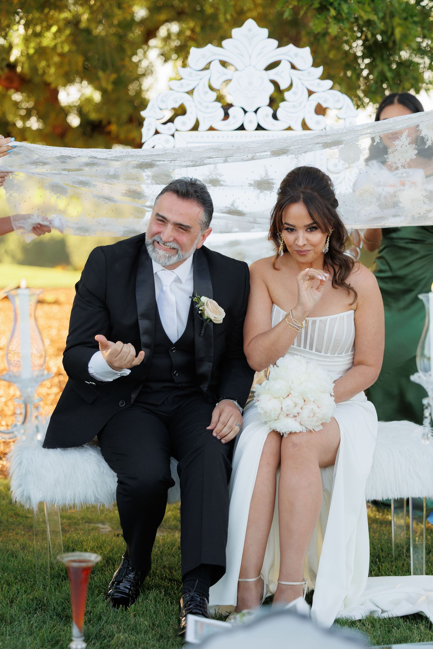 A bride and groom share playful moments while sitting together outdoors near decorative white lattice during their wedding.
