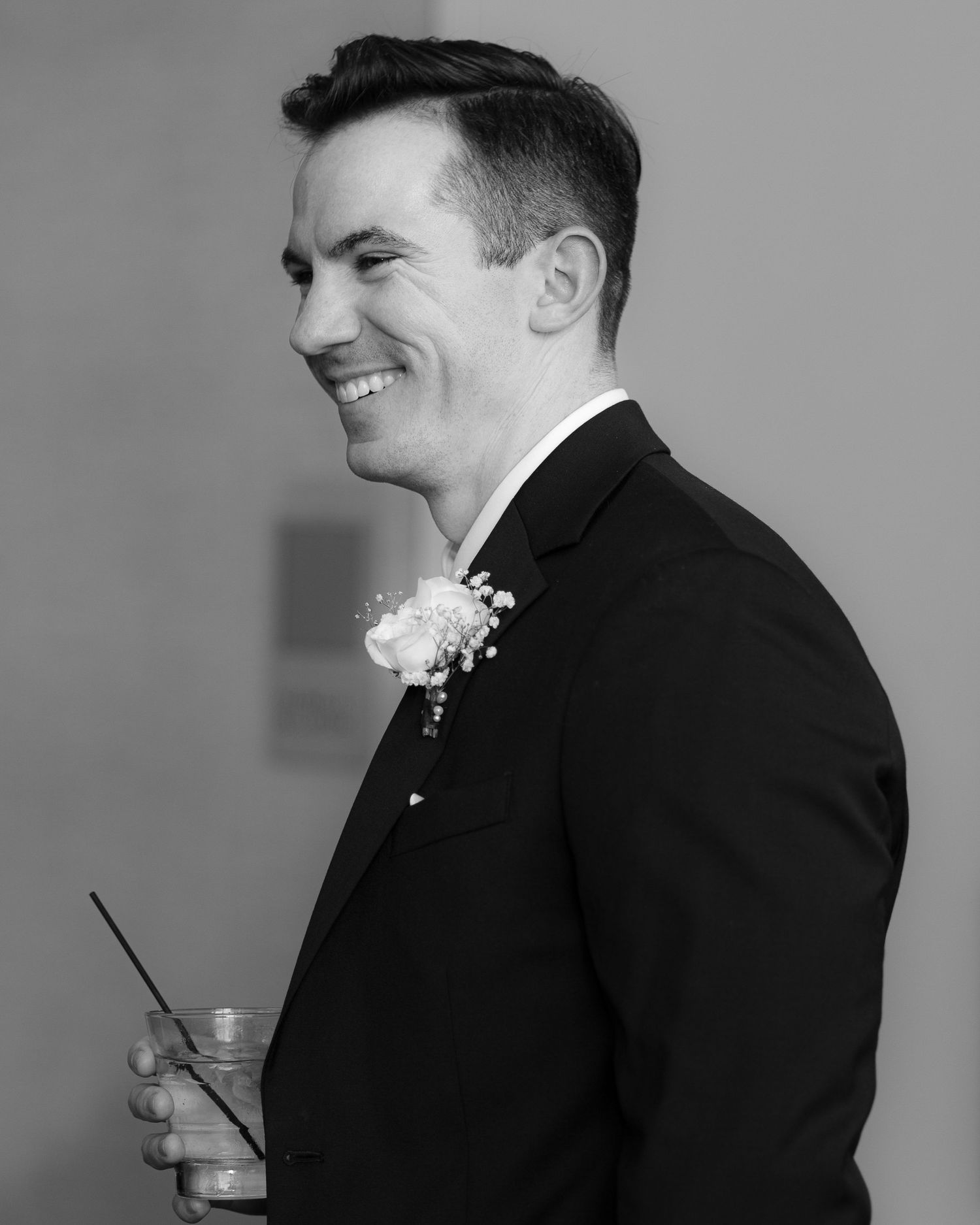 A person in a black suit with a boutonniere smiles while holding a drink in a black and white photograph.