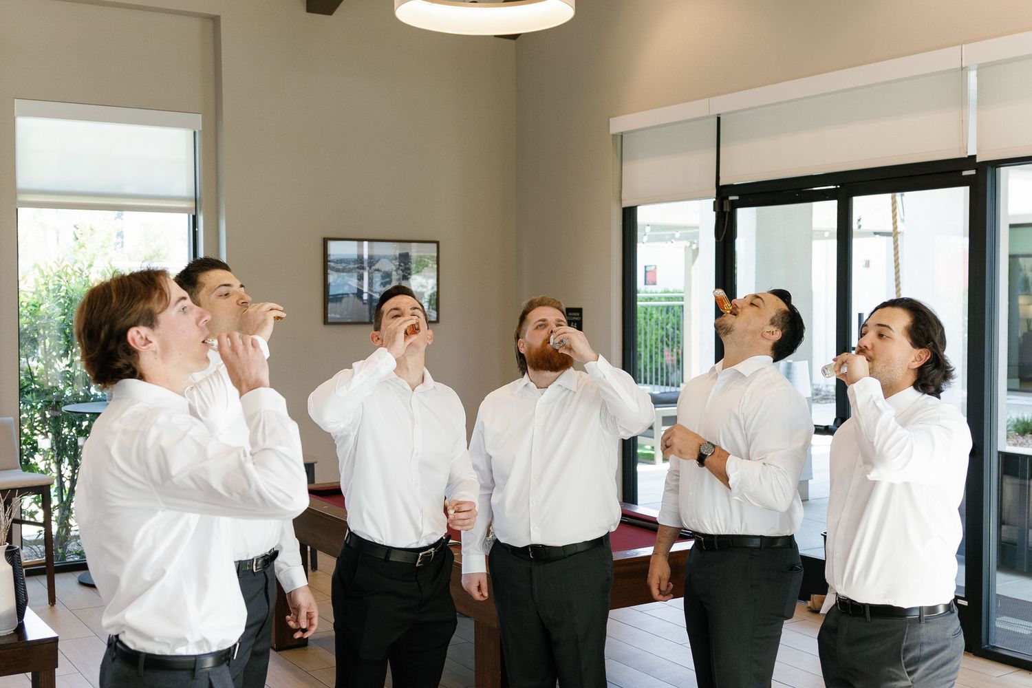 A group of groomsmen in white shirts look up and laugh together in a bright indoor space.