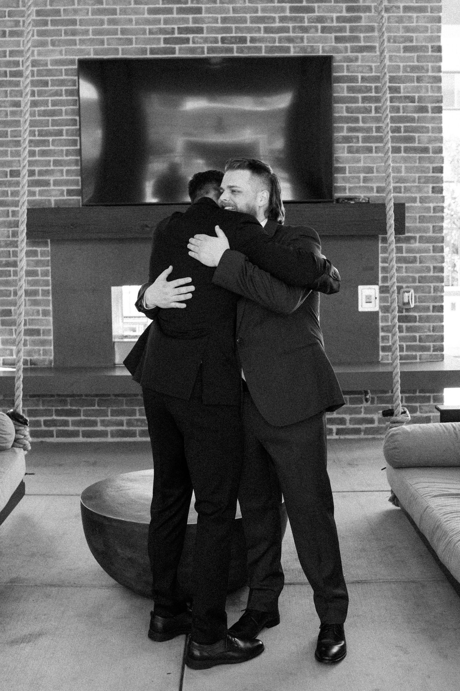 Two people in formal black suits share an emotional embrace in front of a brick wall with a dark display panel.