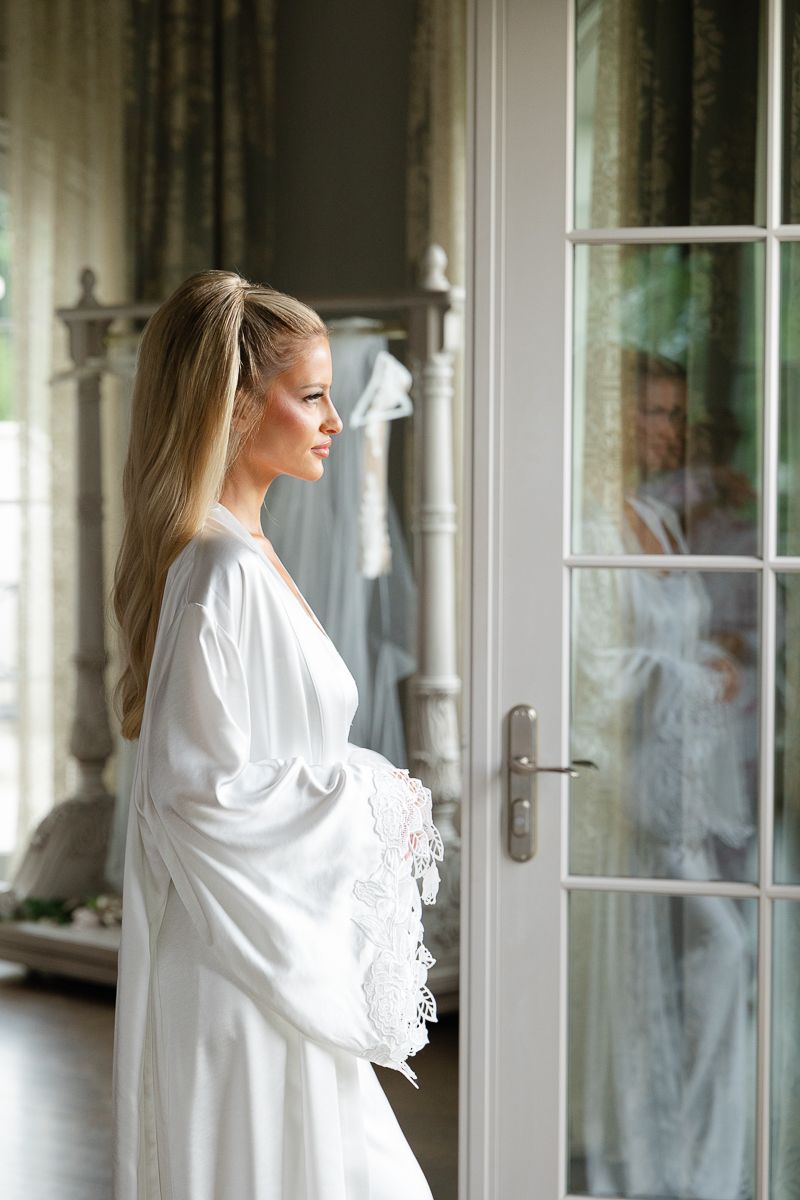 A figure in a white silk robe stands by French doors in a sunlit room with elegant decor.