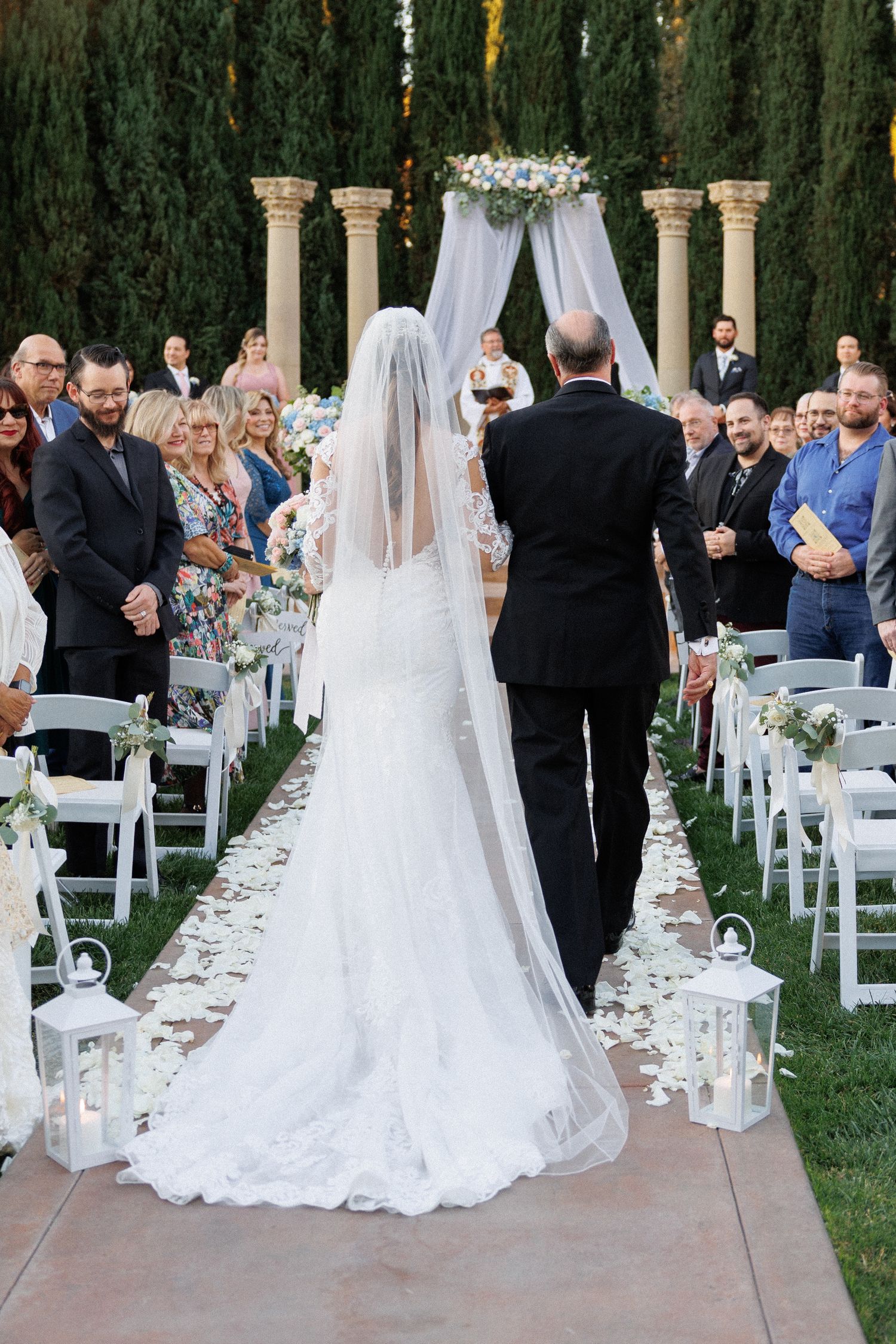 A wedding ceremony procession on a petal-strewn aisle with white lanterns and cypress trees in the background.
