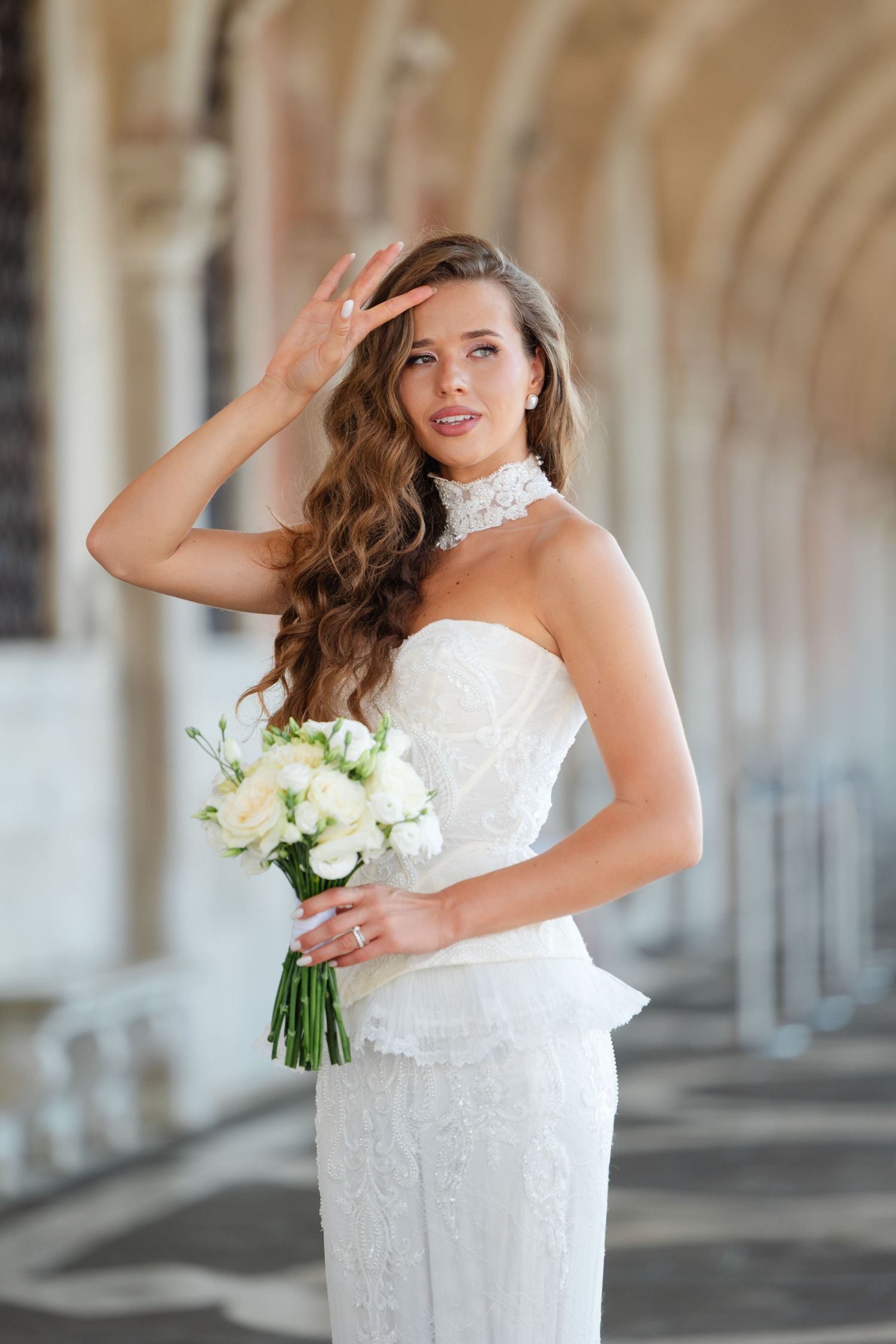 A bride in a fitted white lace gown with high neck detail holds a white bouquet while standing in an arched corridor.