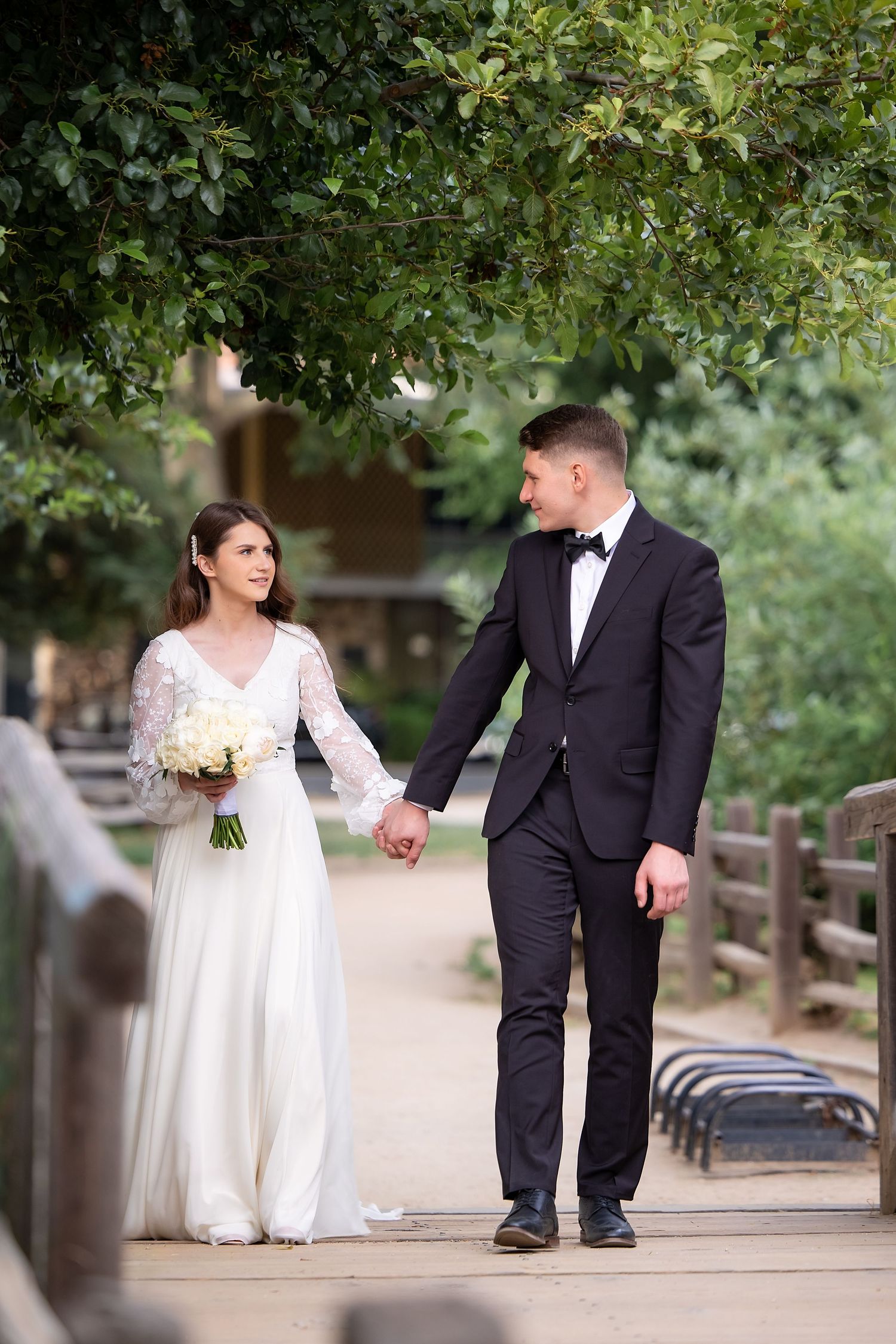 Couple in wedding attire walks hand in hand along garden path surrounded by greenery.