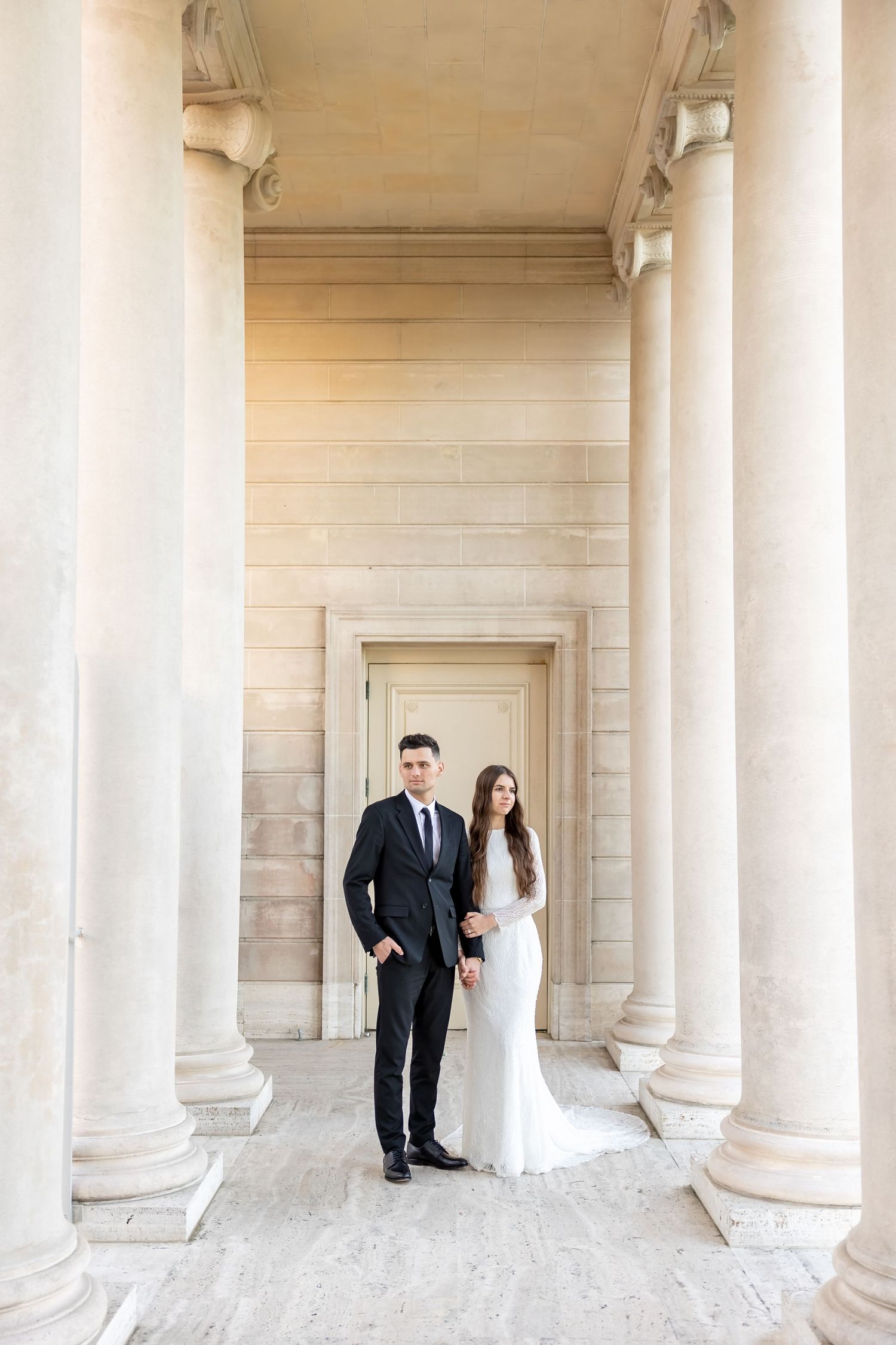 Elegant couple posing between classical columns in formal wedding attire.