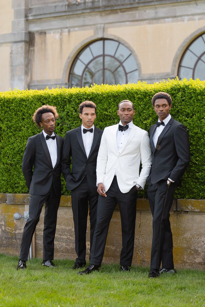 Four men in formal tuxedos and bow ties pose elegantly against a manicured hedge and arched window backdrop.