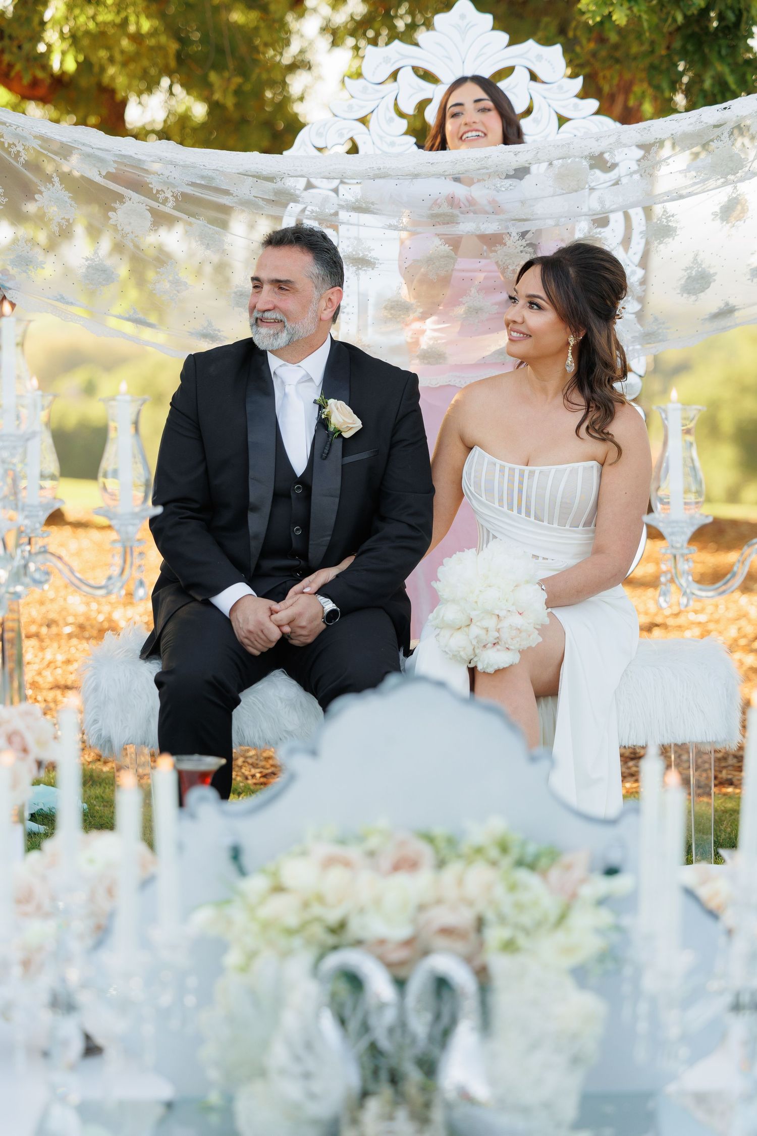 A romantic outdoor wedding scene with a couple sitting at a decorated table surrounded by autumn foliage and white decor.