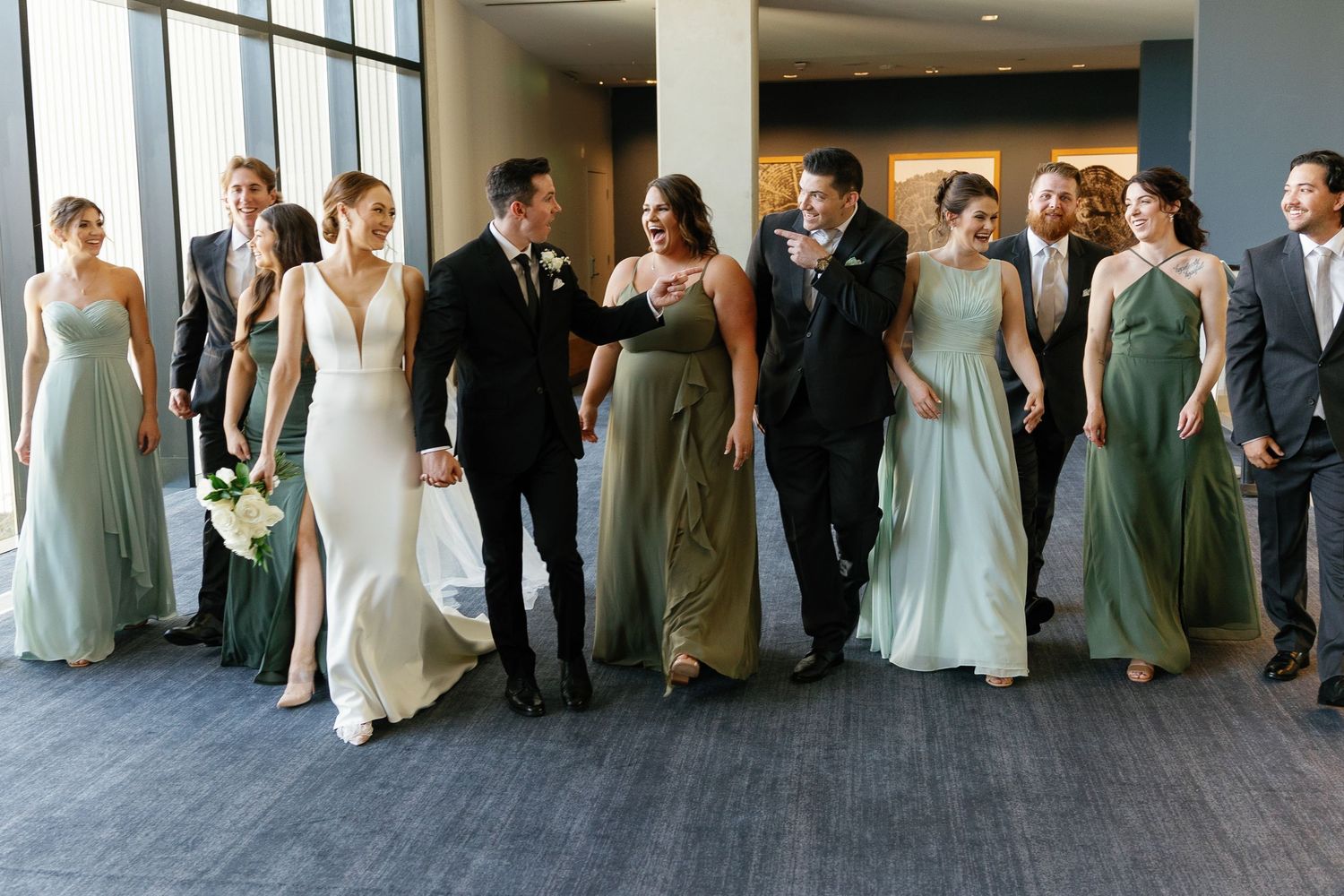 Wedding party members in coordinated sage green and ivory formal attire walk together in an elegant indoor venue.