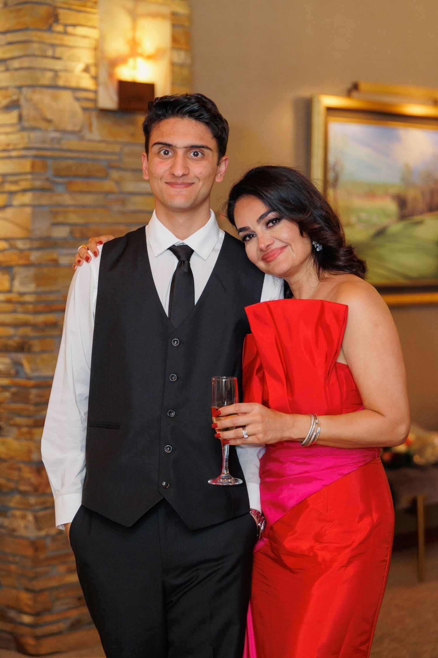 A couple in formal attire poses together at an event with stone walls and artwork in the background.