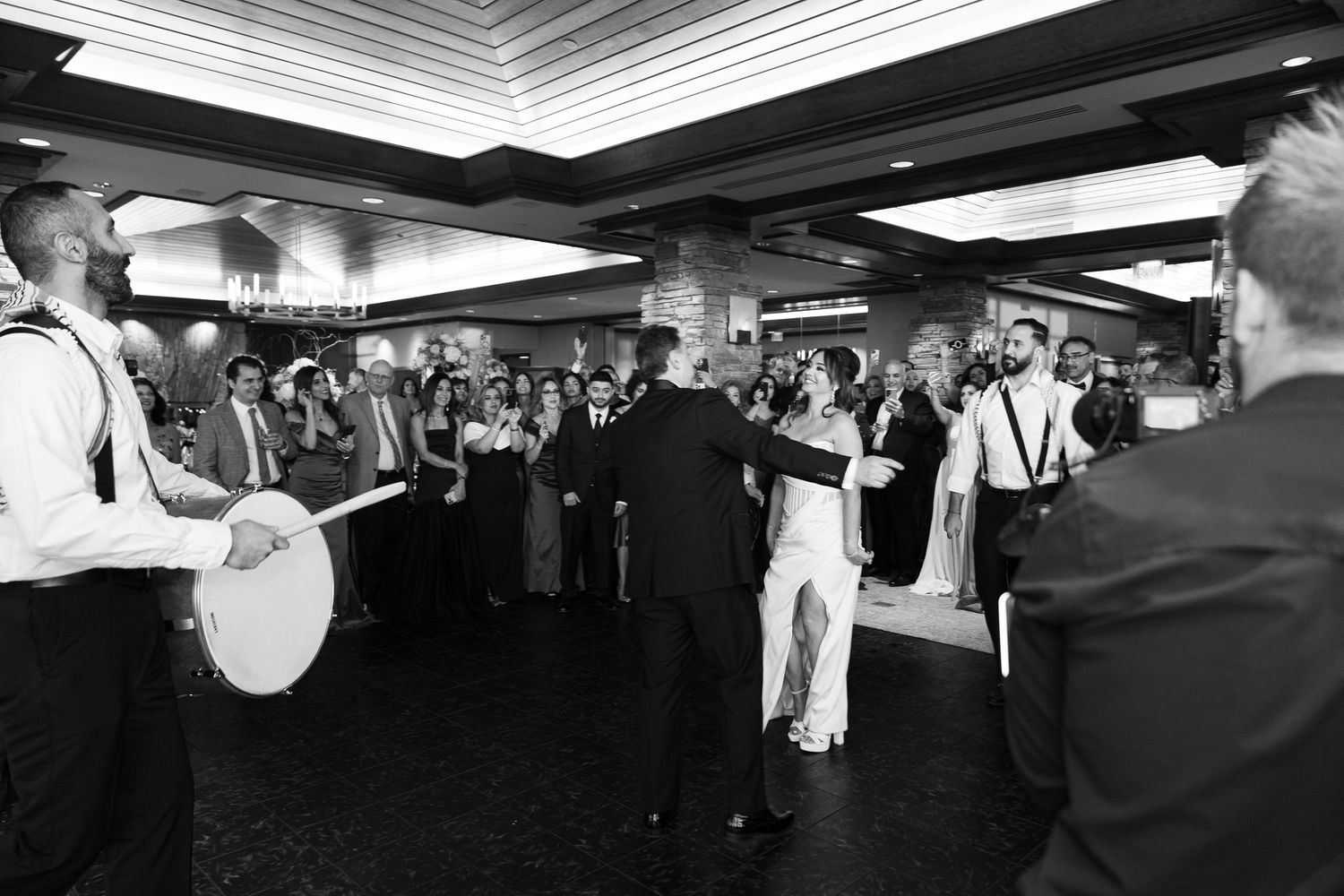 Black and white photo of a lively wedding reception with guests dancing while musicians perform in an indoor venue.