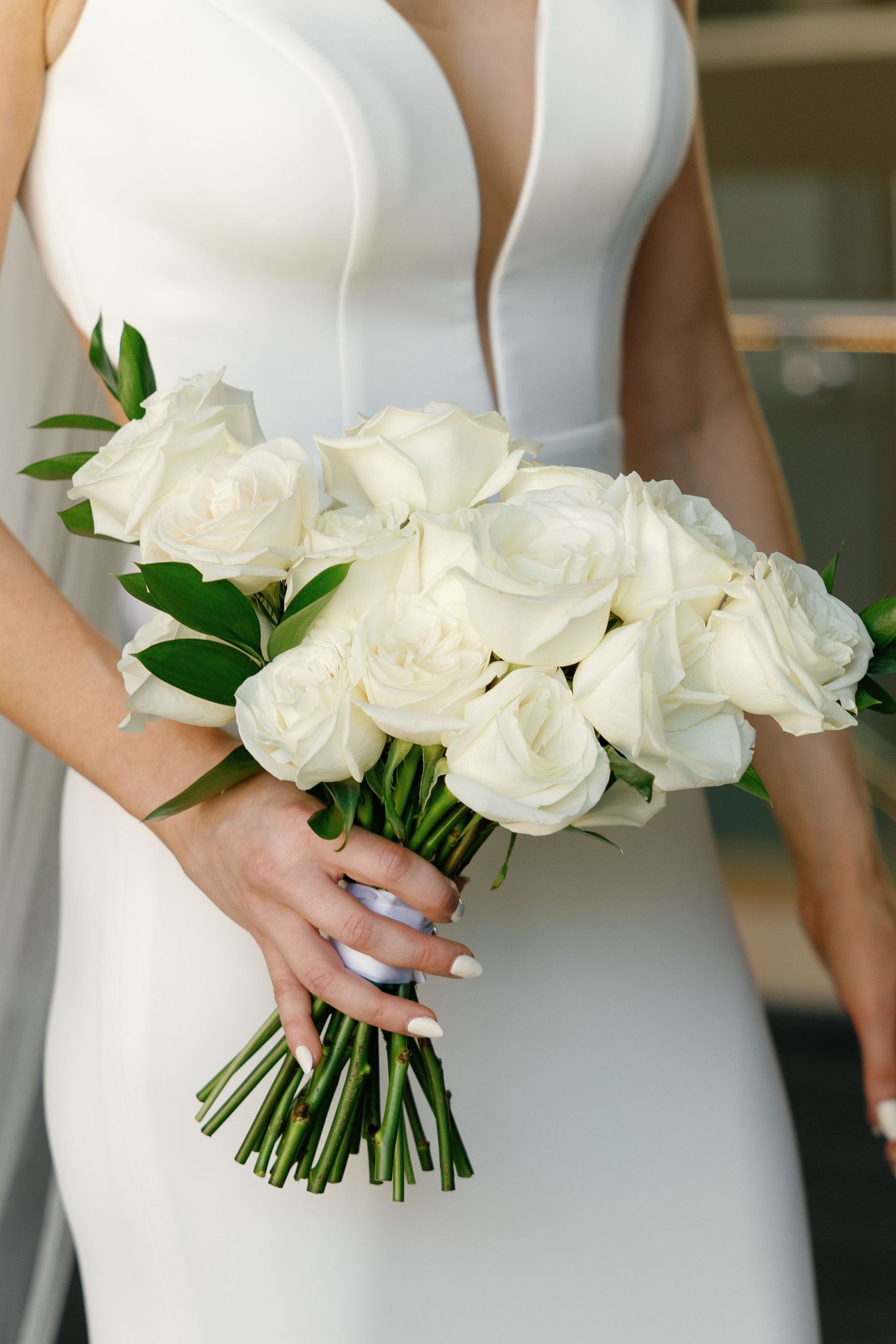 Elegant white bridal bouquet of fresh roses with green foliage held against a fitted white wedding dress.