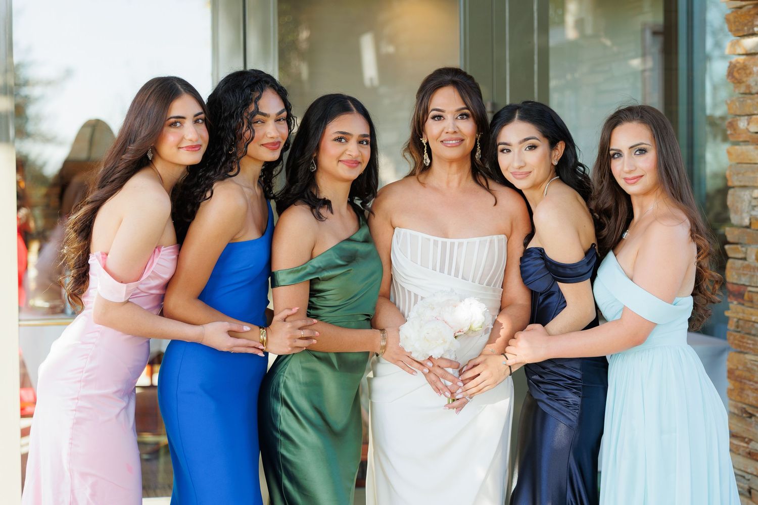 Group of bridesmaids in blue and pastel dresses pose together at a wedding venue.