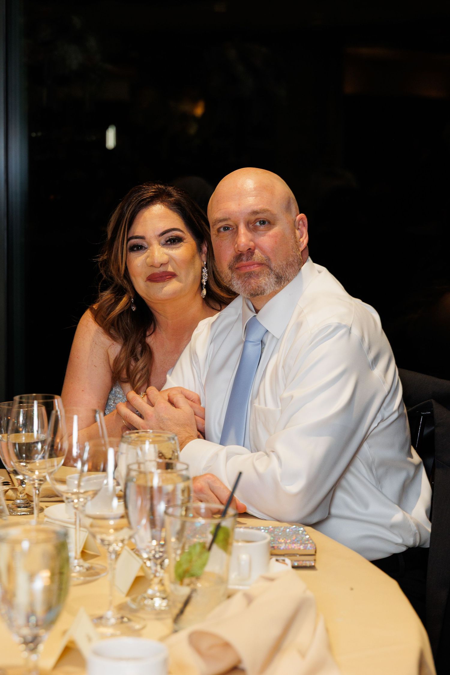 A couple sits together at an elegant dinner table with wine glasses and place settings in a dimly lit restaurant setting.