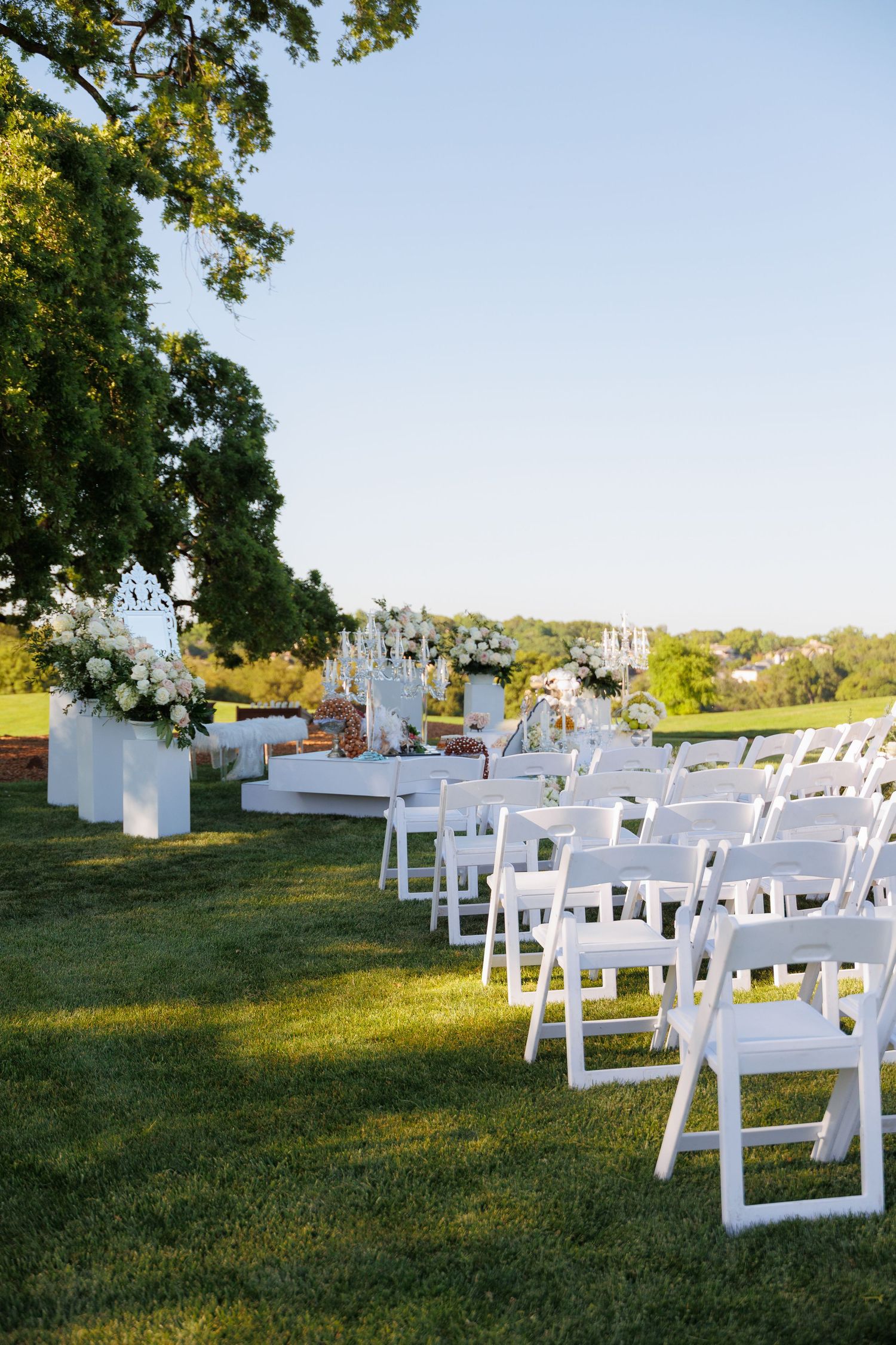 White wooden folding chairs arranged in rows on a grassy lawn for an outdoor ceremony under shady trees.