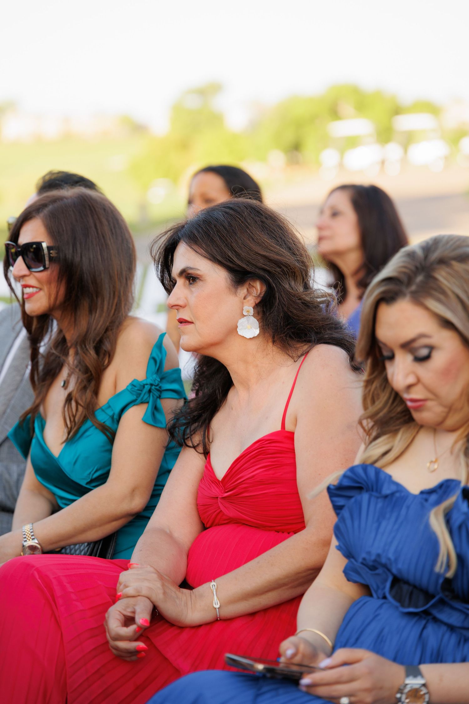 A group of women in colorful dresses sit together outdoors wearing sunglasses and enjoying a sunny day.
