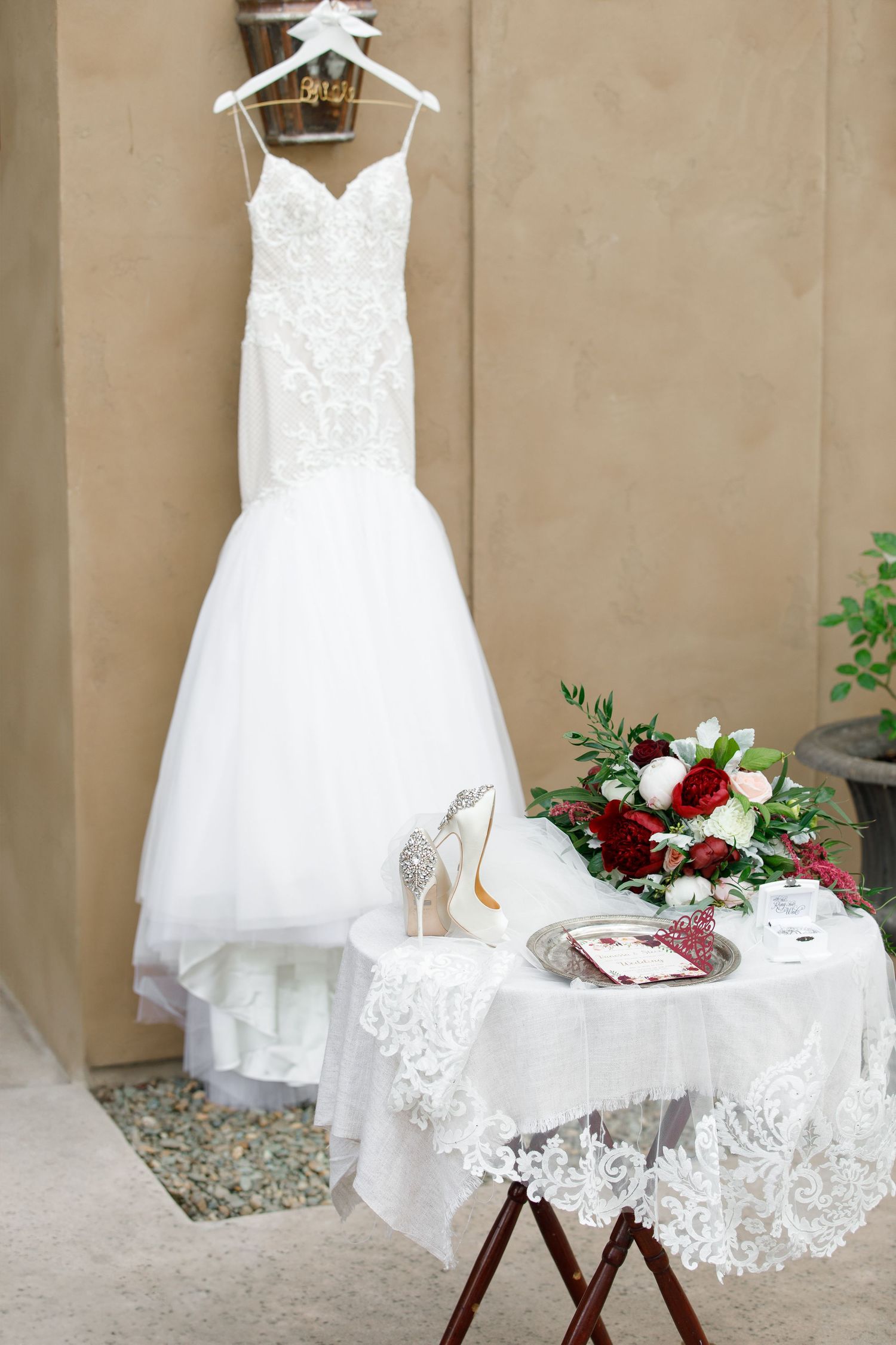 A white lace wedding dress hangs elegantly on a hanger against a beige wall with decorative table setting below.