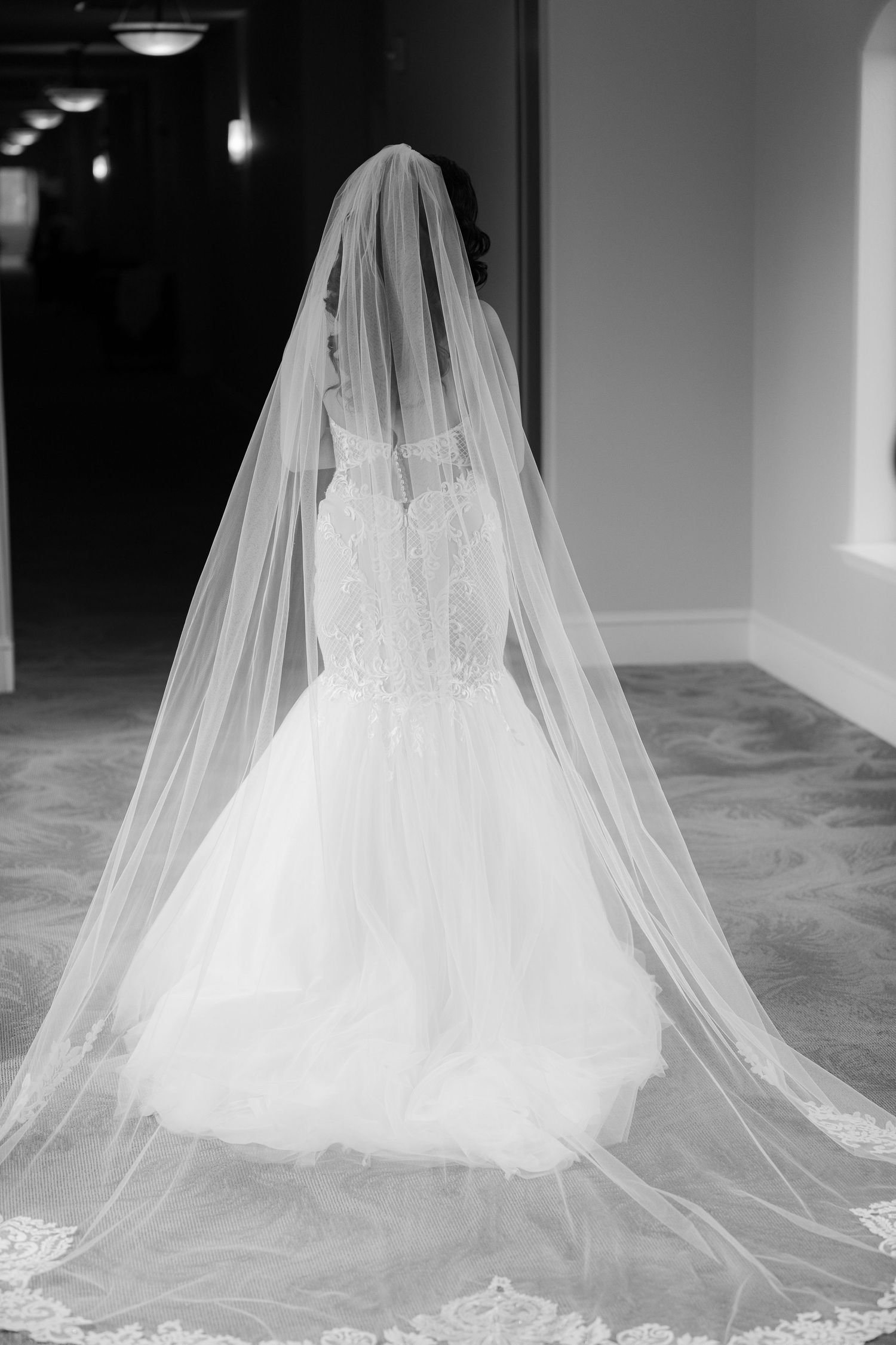 A dramatic black and white photograph of a wedding dress with flowing cathedral veil on a light colored floor.