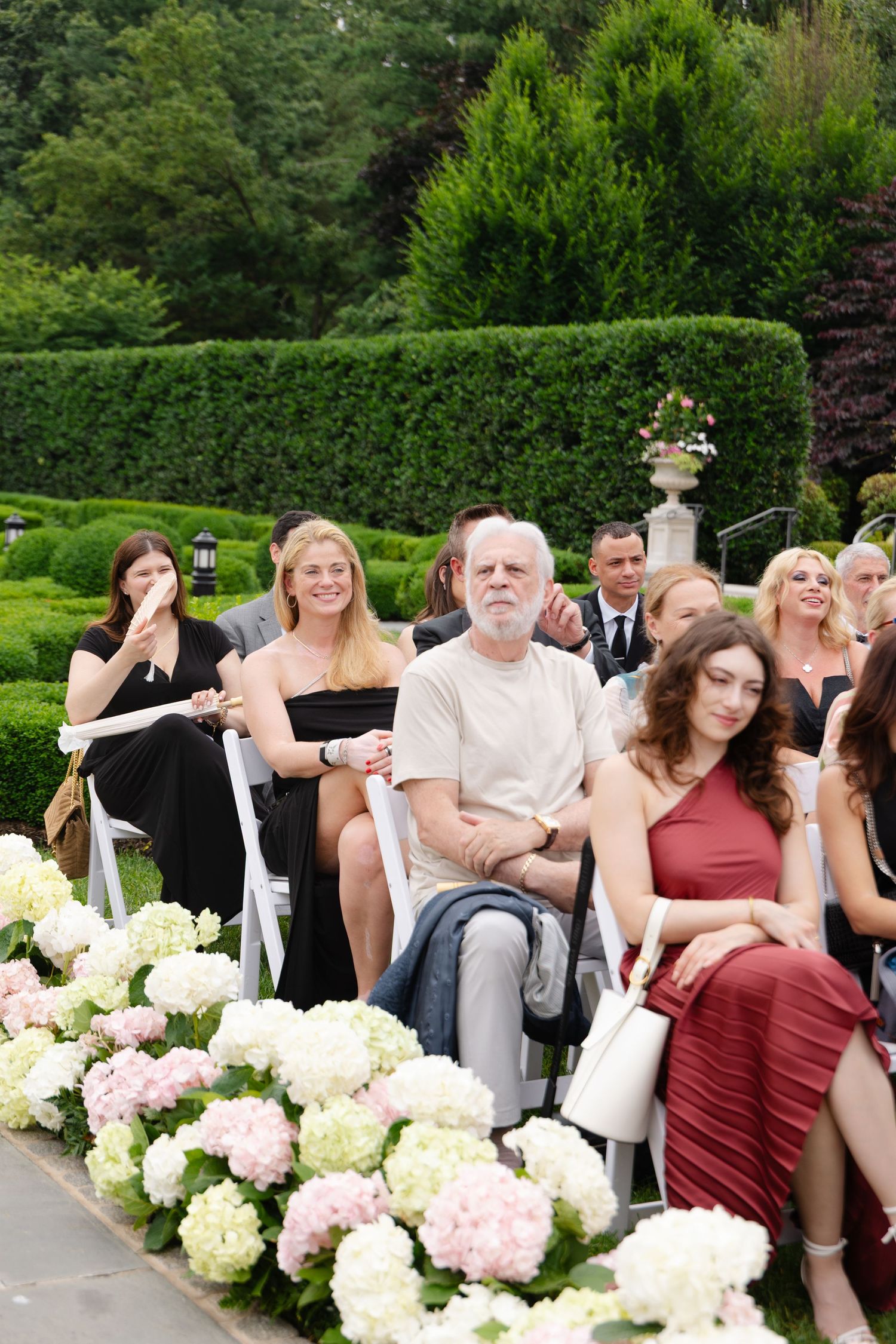 Wedding guests seated in formal garden setting with lush hedges and hydrangea floral arrangements along the aisle.