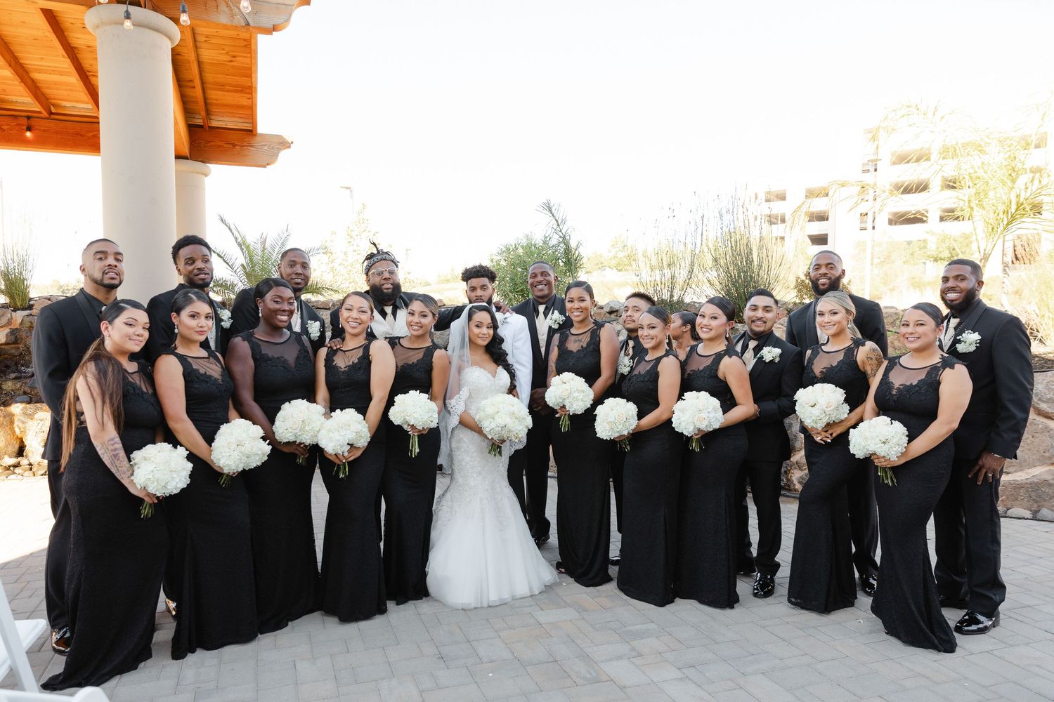Wedding party in black and white attire poses together in outdoor courtyard for formal group photos.