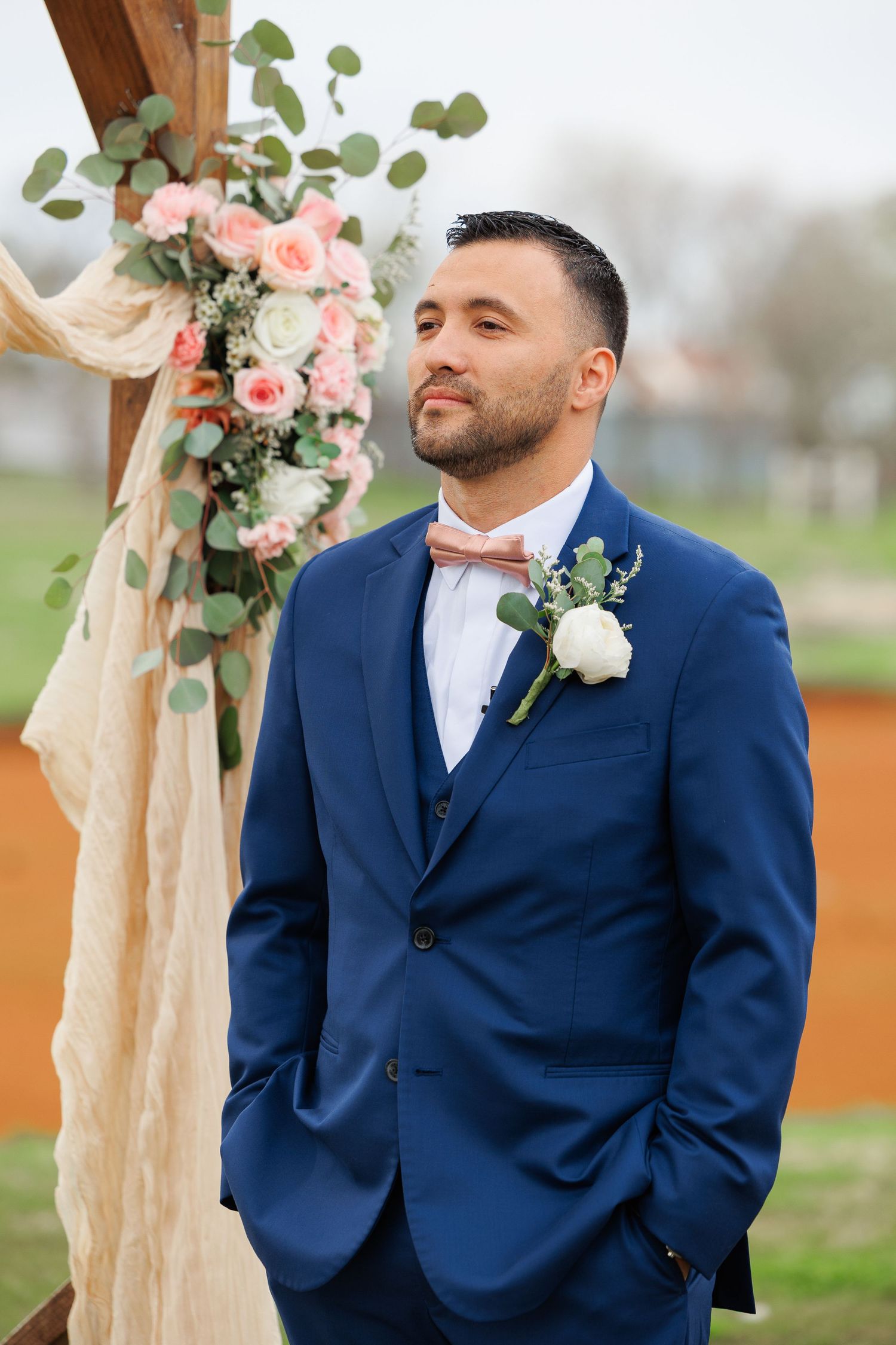 Wedding attire features a navy blue suit with white boutonniere and bow tie against floral arch decorations.
