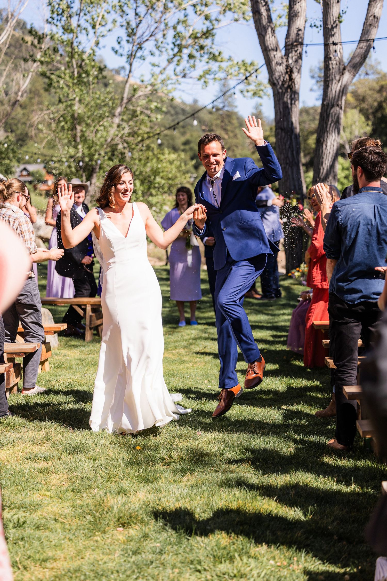 Wedding guests dance and celebrate outdoors on a grassy lawn surrounded by trees on a sunny afternoon.