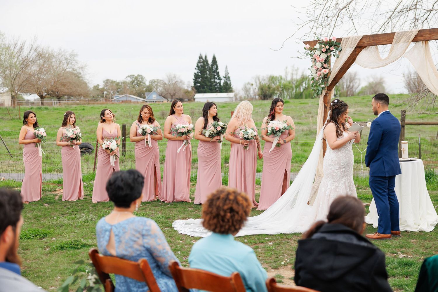 A wedding party celebrates outdoors in a spring field with bridesmaids wearing pink dresses and a bride in white.