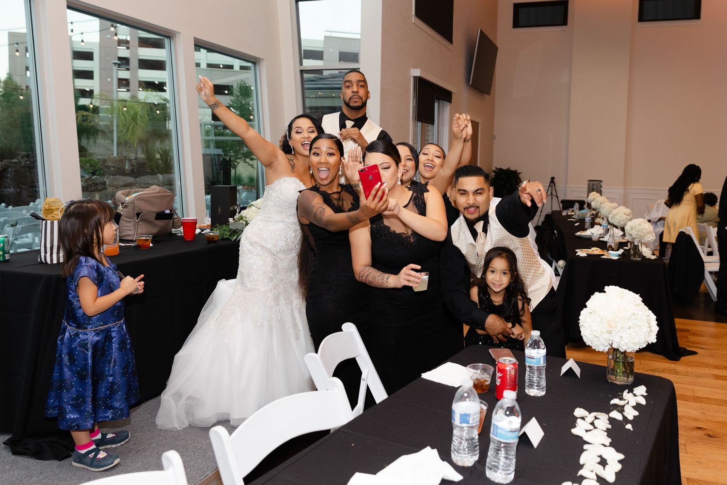 People dancing and celebrating at a wedding reception in an indoor venue with large windows.