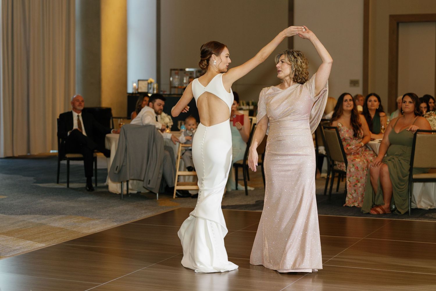 Two people in elegant dresses share a dance together on a ballroom floor with guests seated in the background.