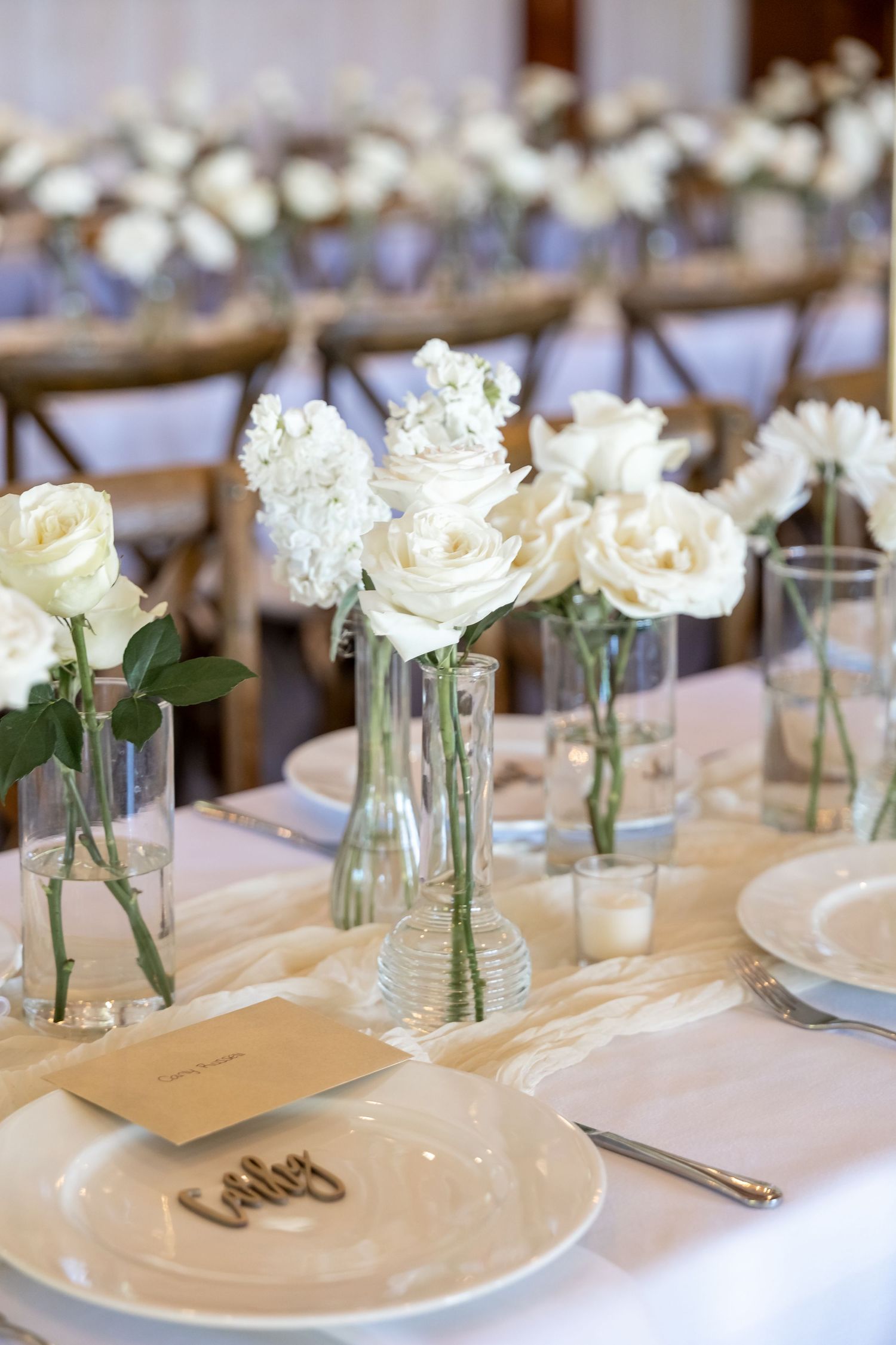 Elegant wedding table setting with white roses in glass vases and white place settings.