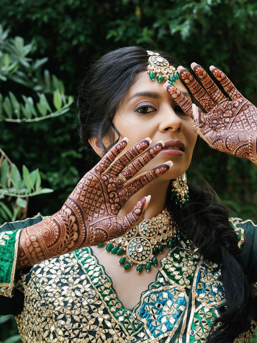 Traditional Indian bridal mehndi design displayed on hands with ornate jewelry and emerald green accessories.