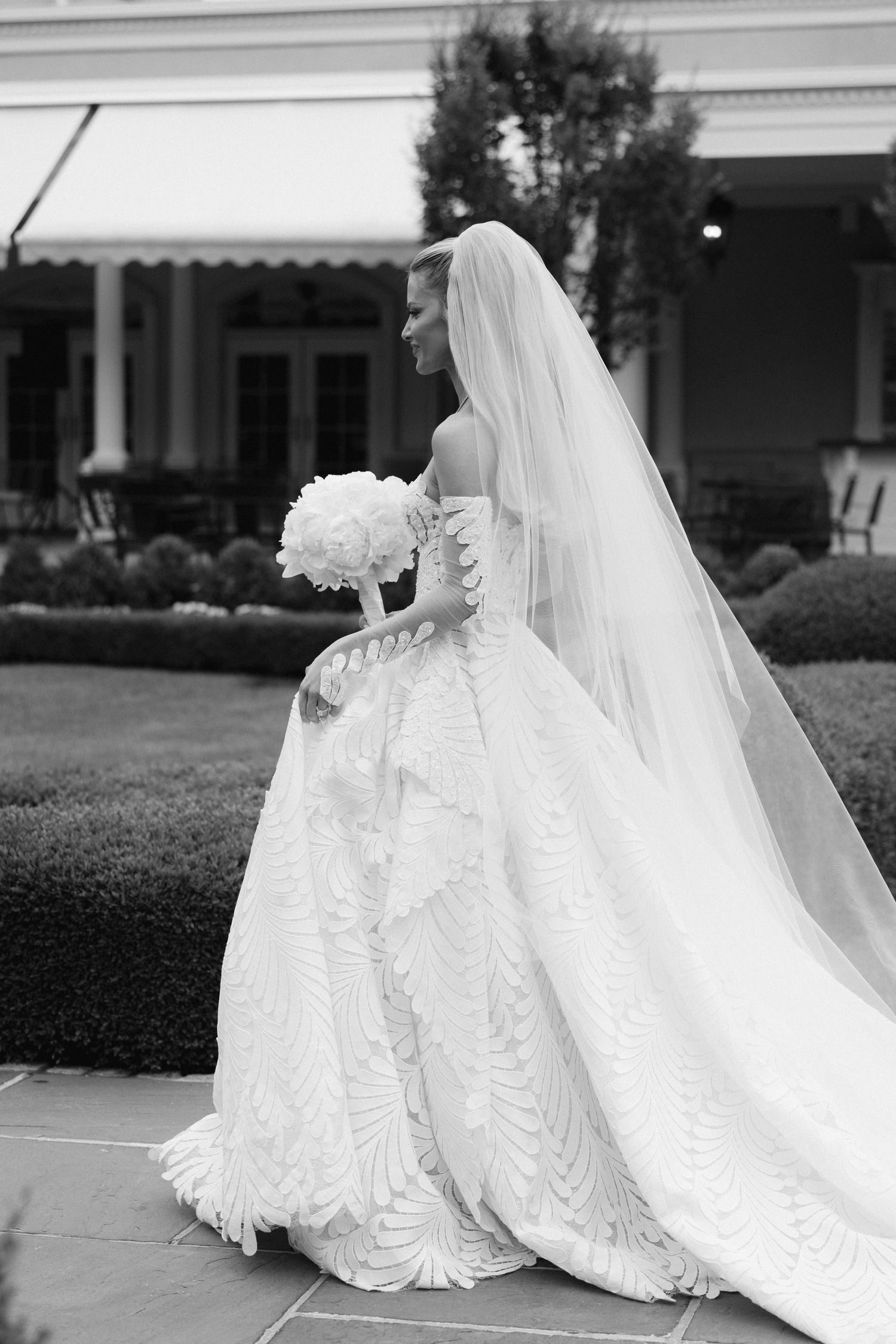 Black and white photo of a bride in an ornate off-shoulder gown with veil, holding a bouquet in an elegant garden.