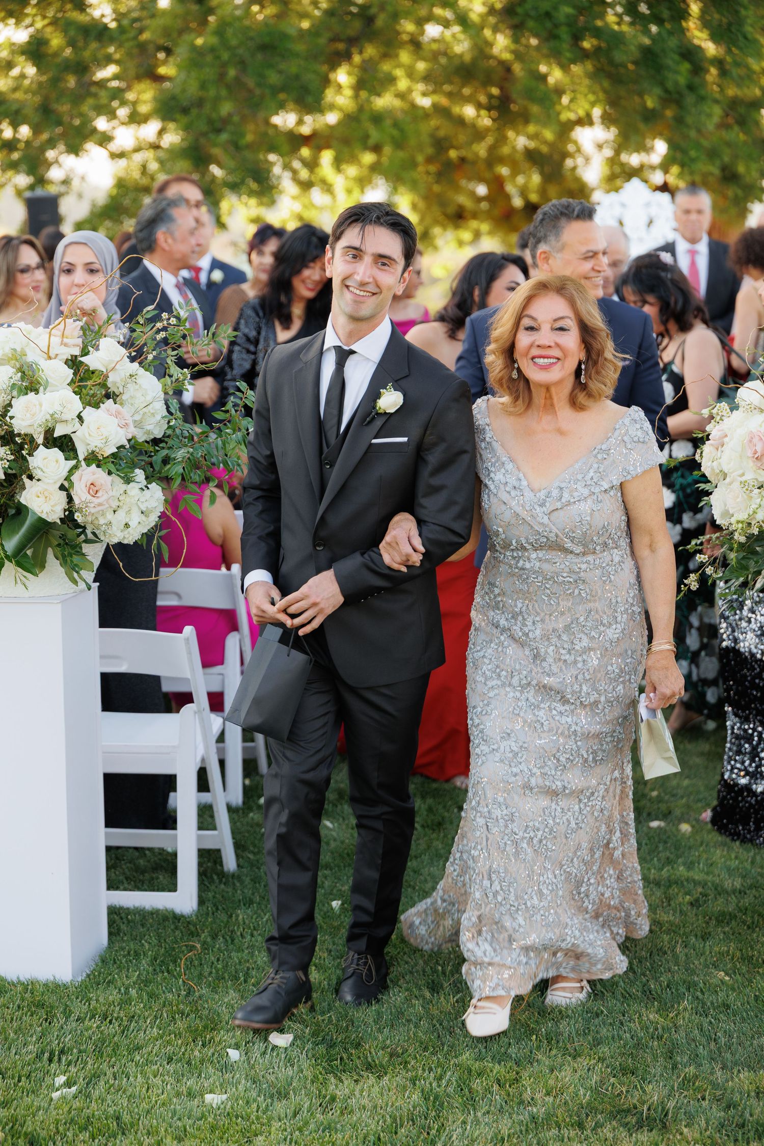 Wedding guests in formal attire walk together on a grassy lawn decorated with white floral arrangements.