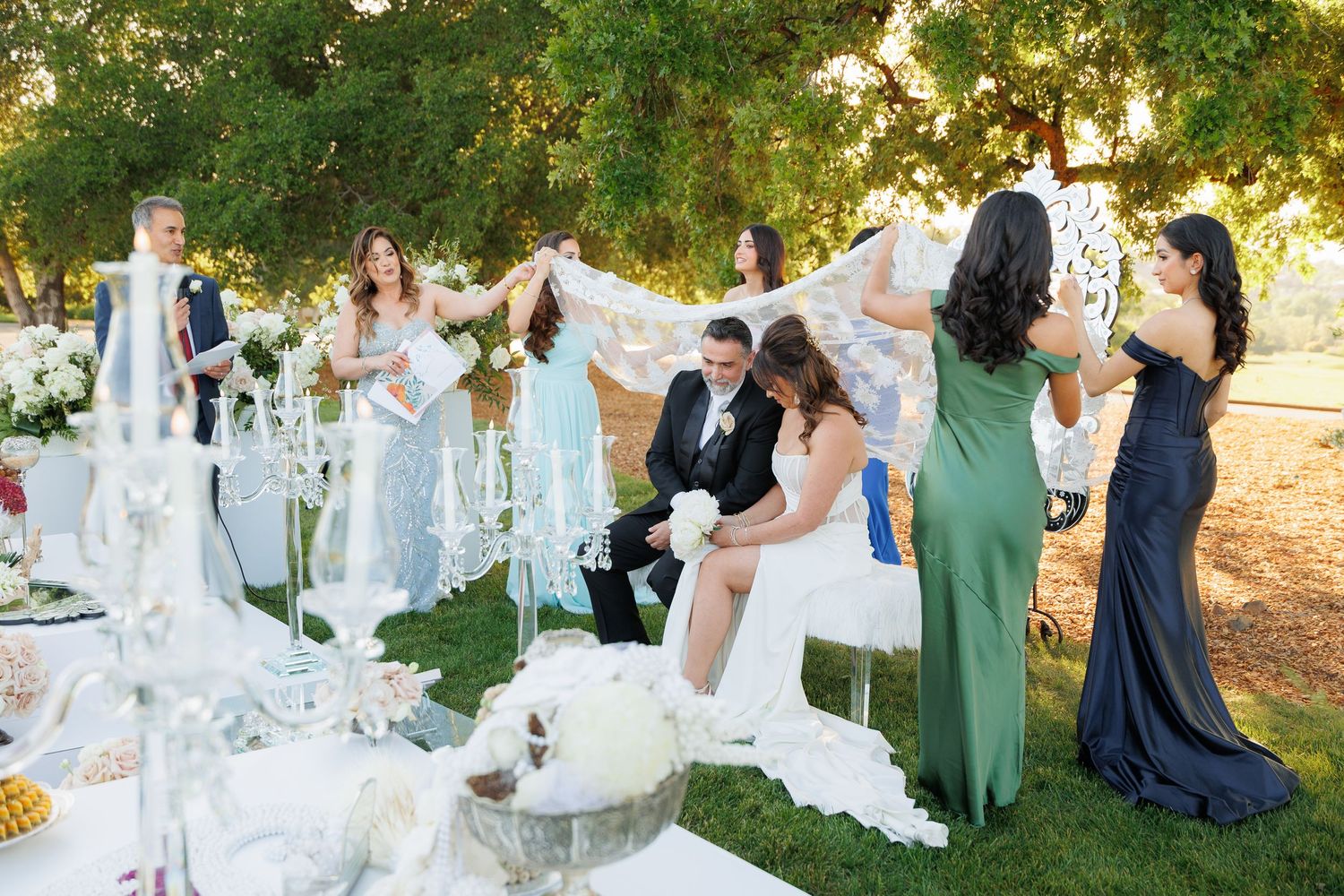 Wedding party celebrates during an elegant outdoor garden reception with white decor and flowing dresses.