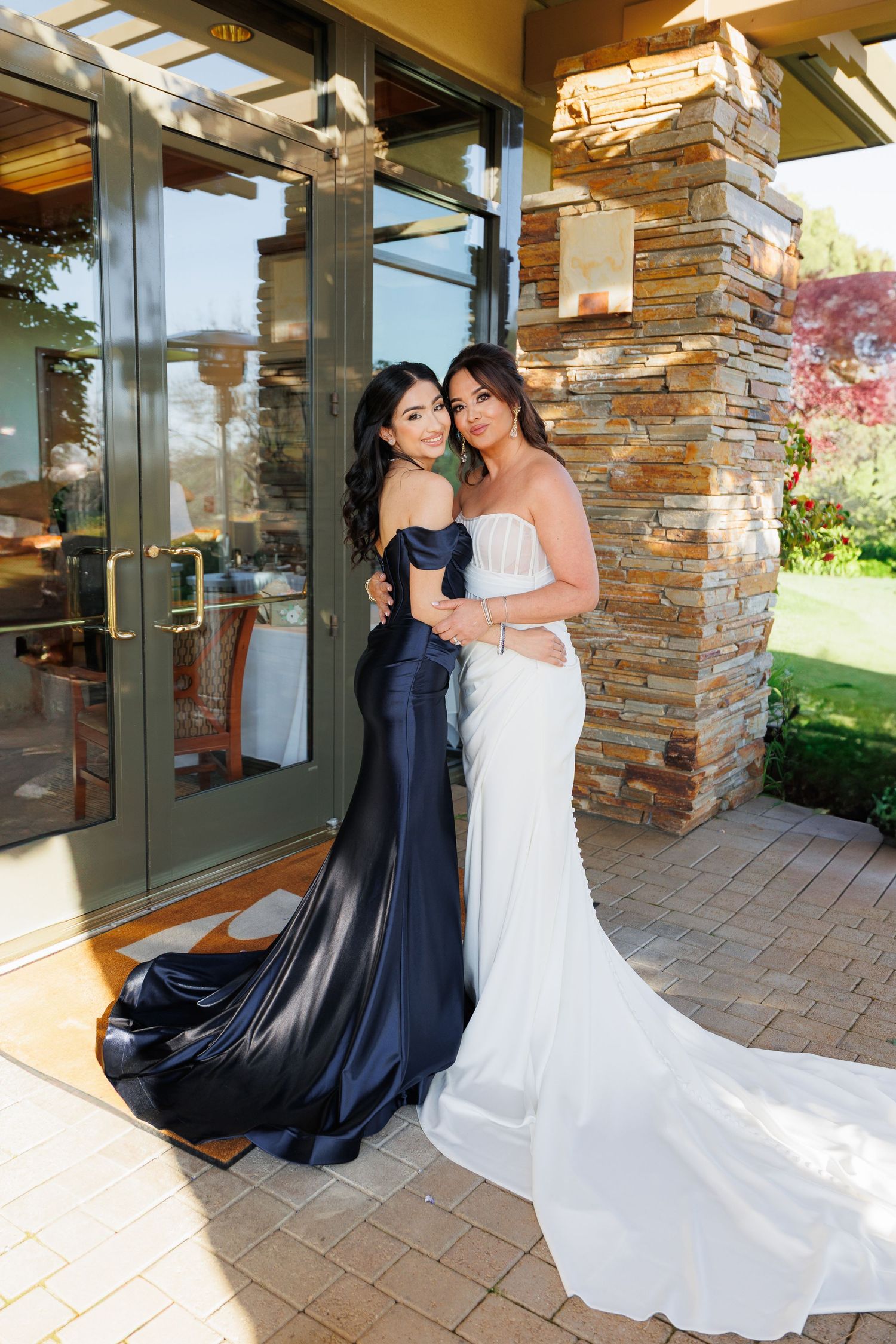 Two figures in contrasting navy and white gowns share an intimate moment on a stone-paved patio at a wedding venue.