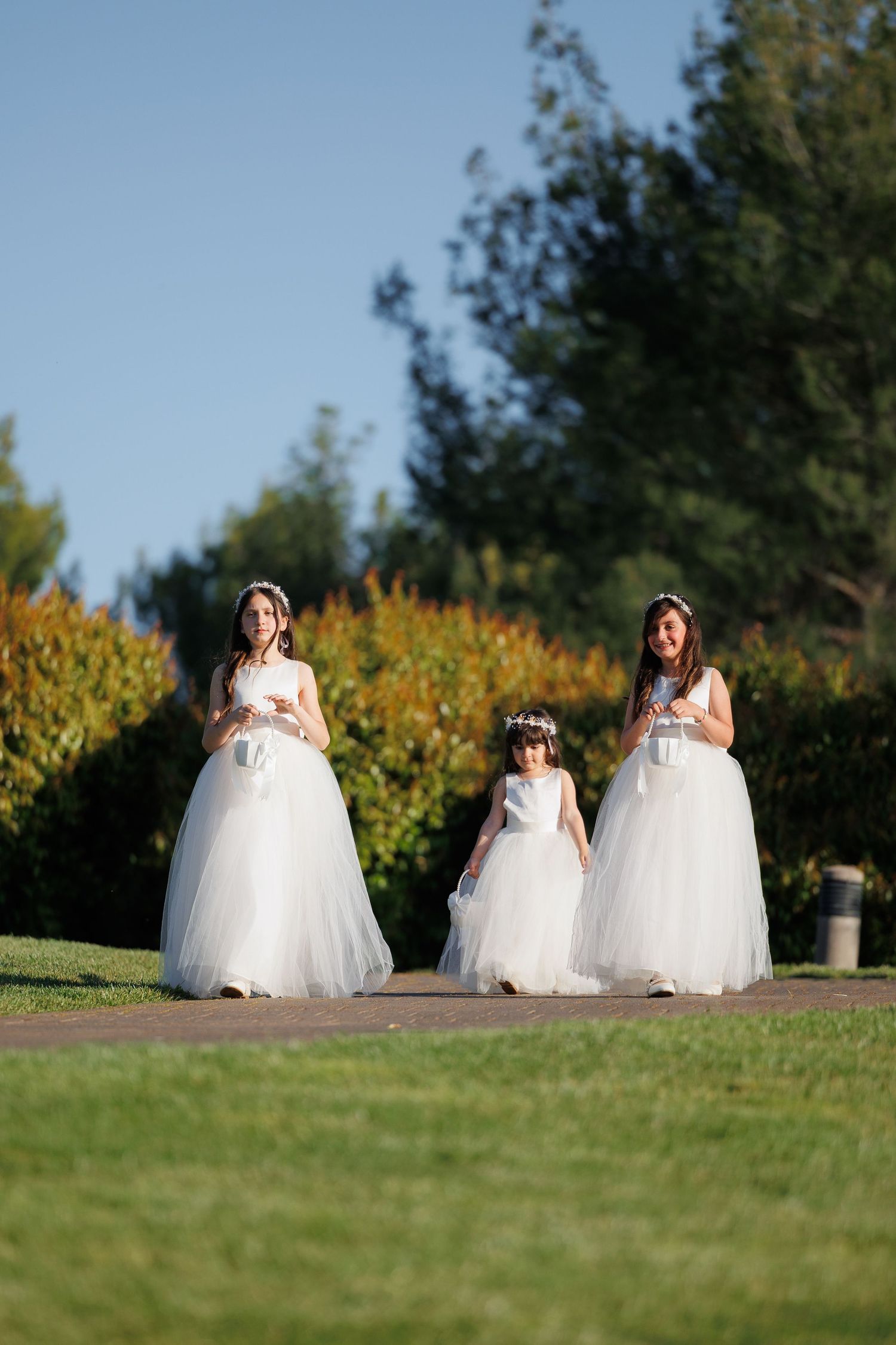 Three figures in flowing white dresses stand together on a grassy lawn with yellow flowers and trees in the background.