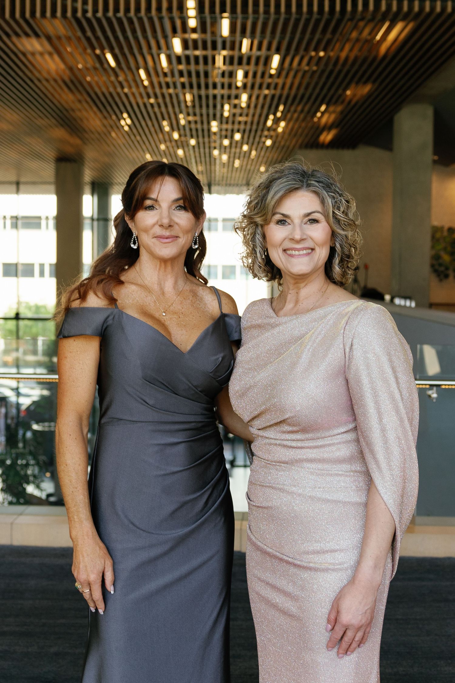 Two women pose together in elegant evening attire, one wearing a charcoal gray dress and one in a shimmering blush gown.