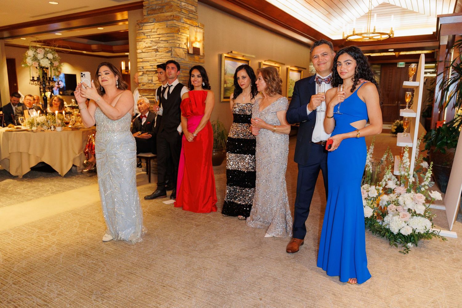 Guests in formal evening attire gather at an elegant reception venue with stone walls and floral decorations.