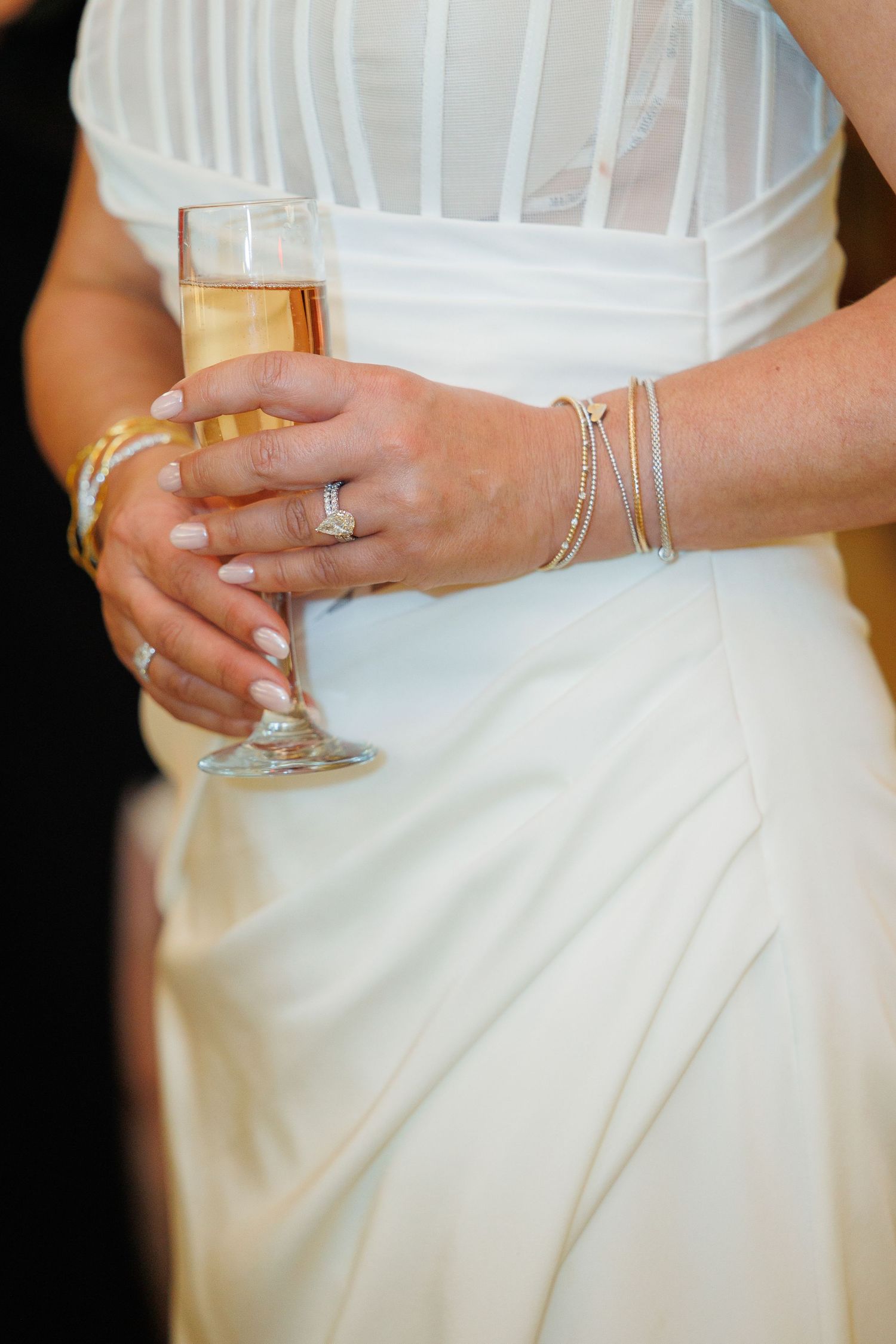 A bride in a white dress holds a champagne glass, showing delicate jewelry on her wrist and hand.