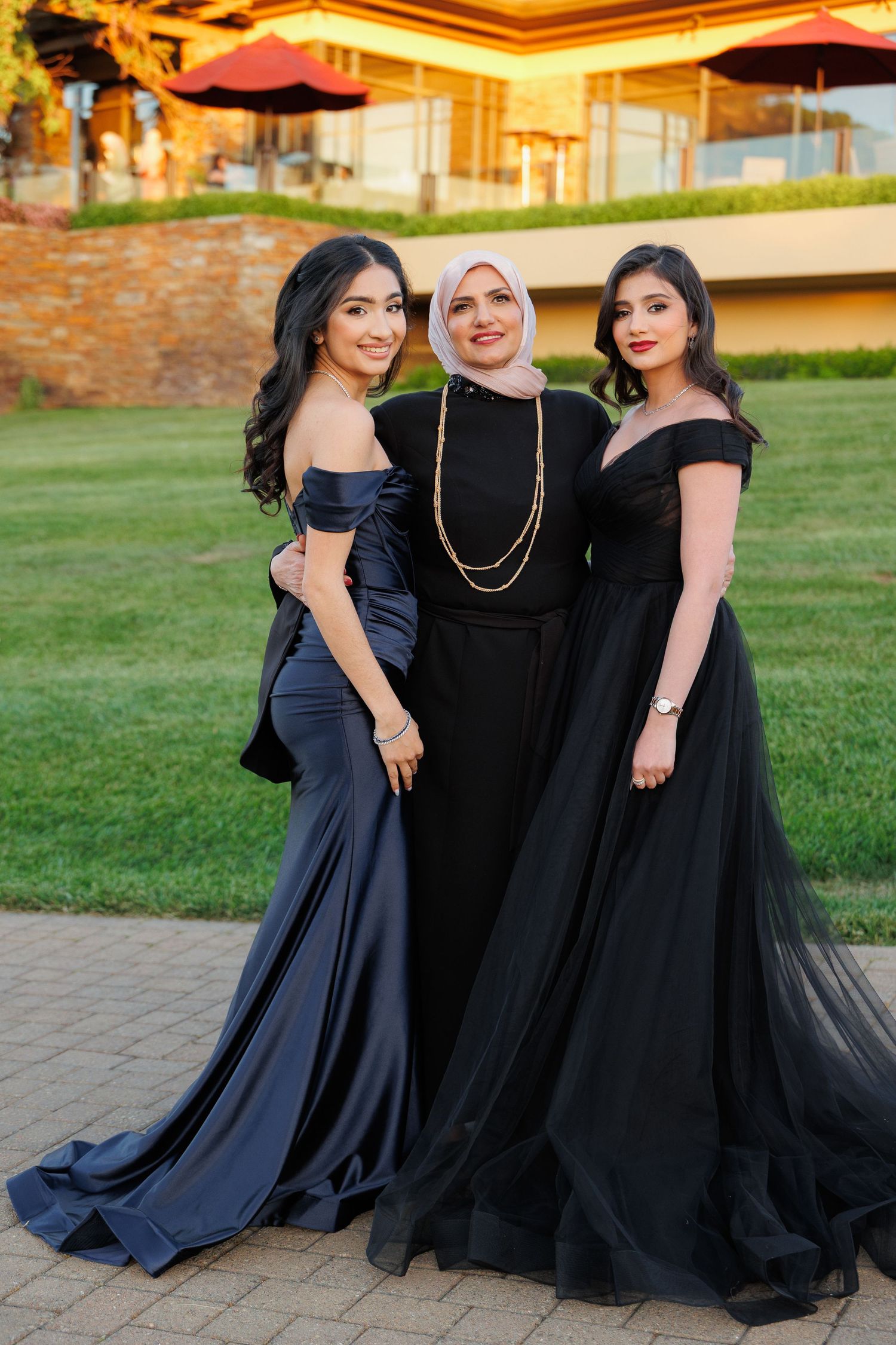 Three friends in elegant evening gowns pose together on a lawn at sunset during a formal event.